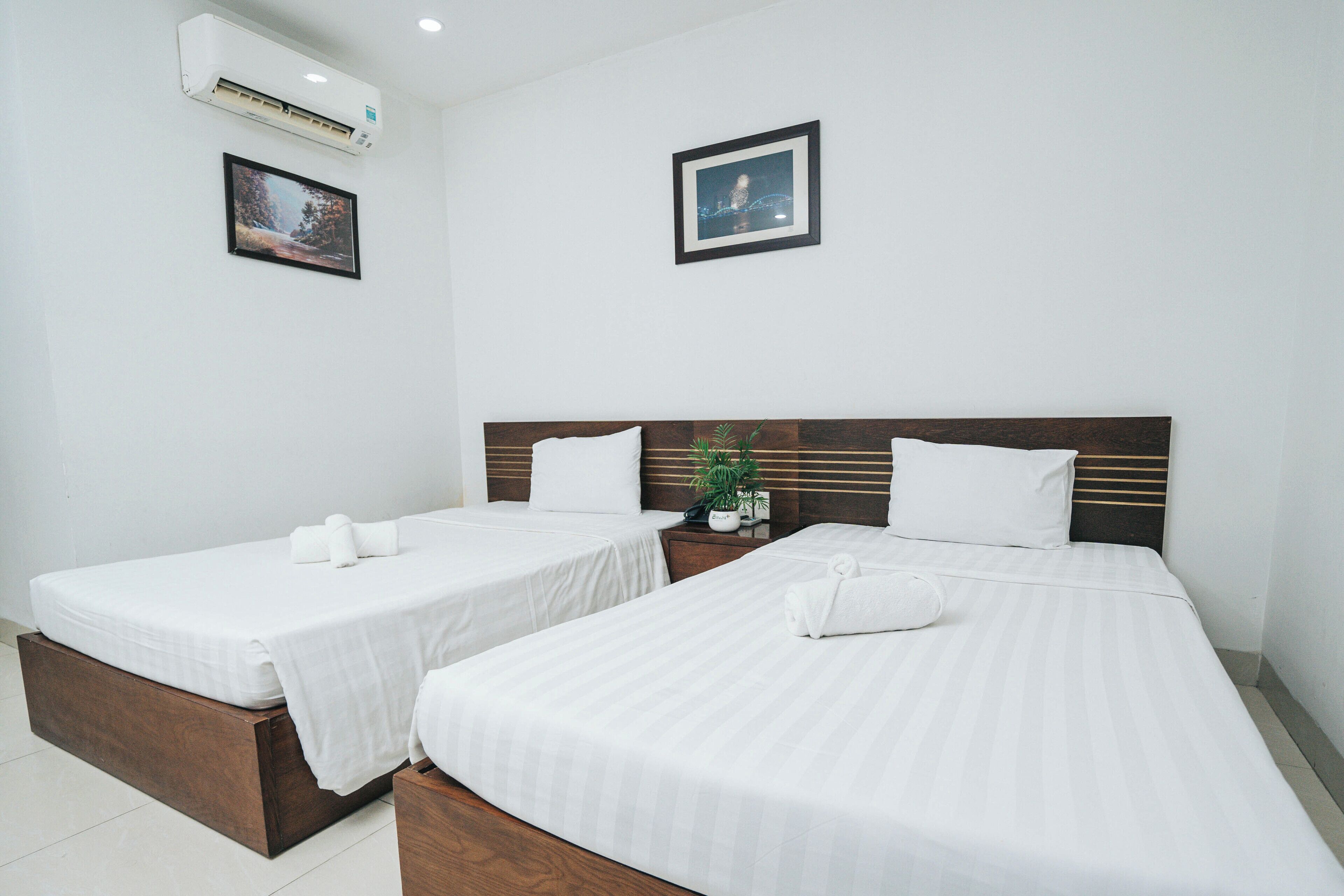 A hotel room with two single beds, a wooden headboard, a small table with a plant, and a framed picture on the wall.