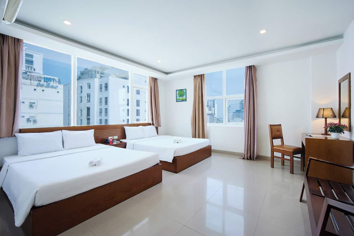 A hotel room with two beds, a desk, a chair, and a view of the cityscape.