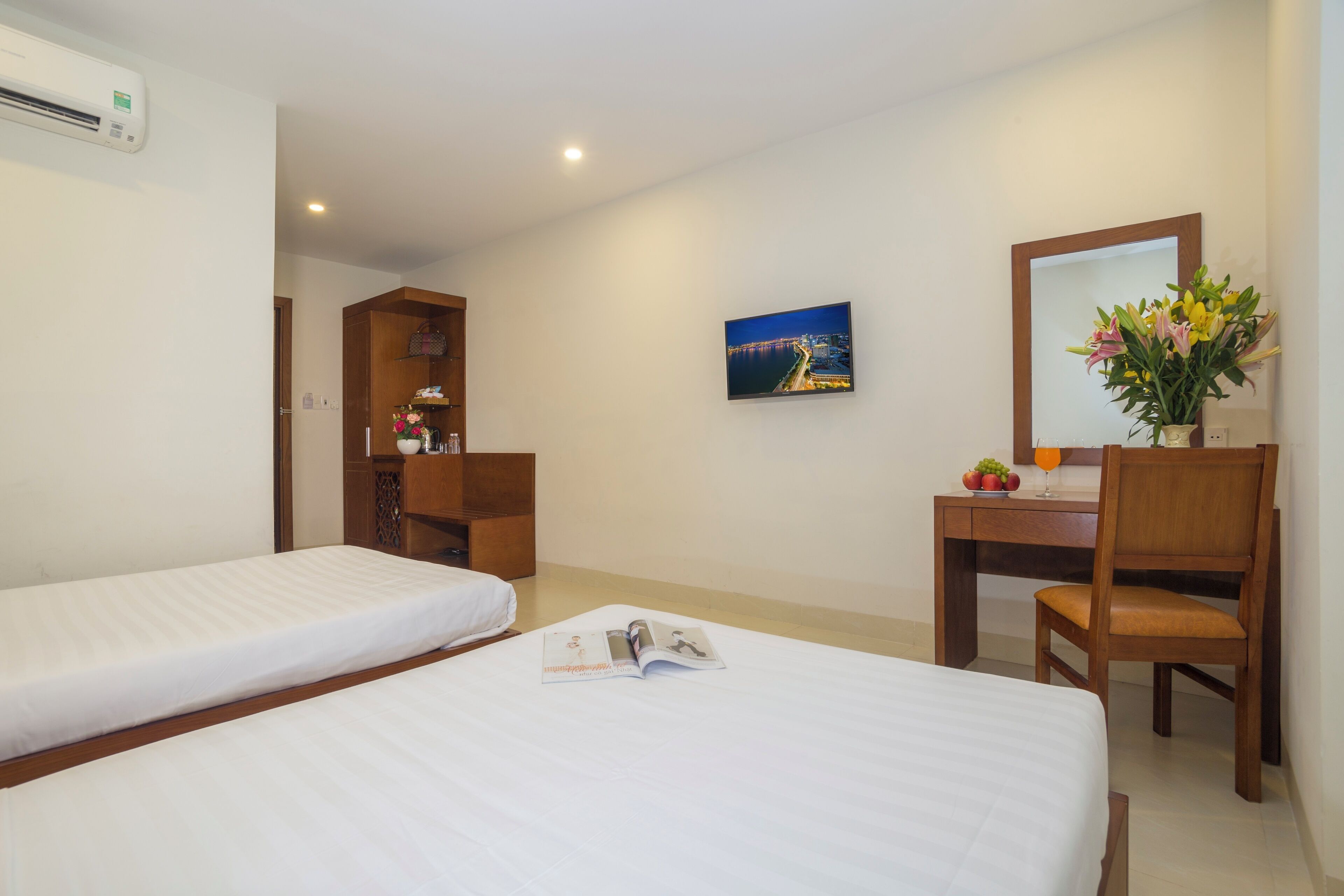 A hotel room with two beds, a desk, a chair, a mirror, a picture on the wall, and a vase with flowers.