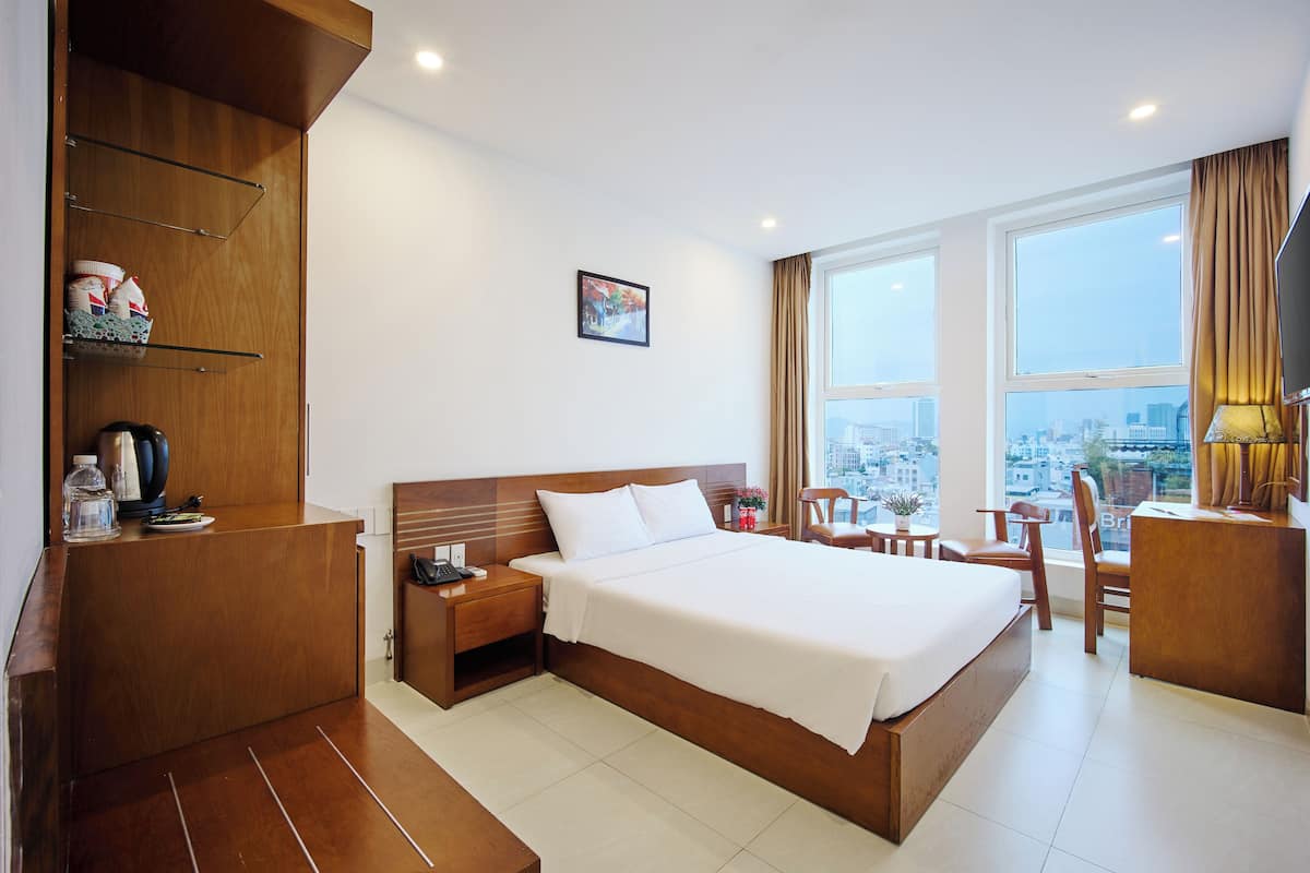 A hotel room with a large bed, wooden headboard, a desk with a chair, a small table with a chair, a window with a city view, and a shelf with items.