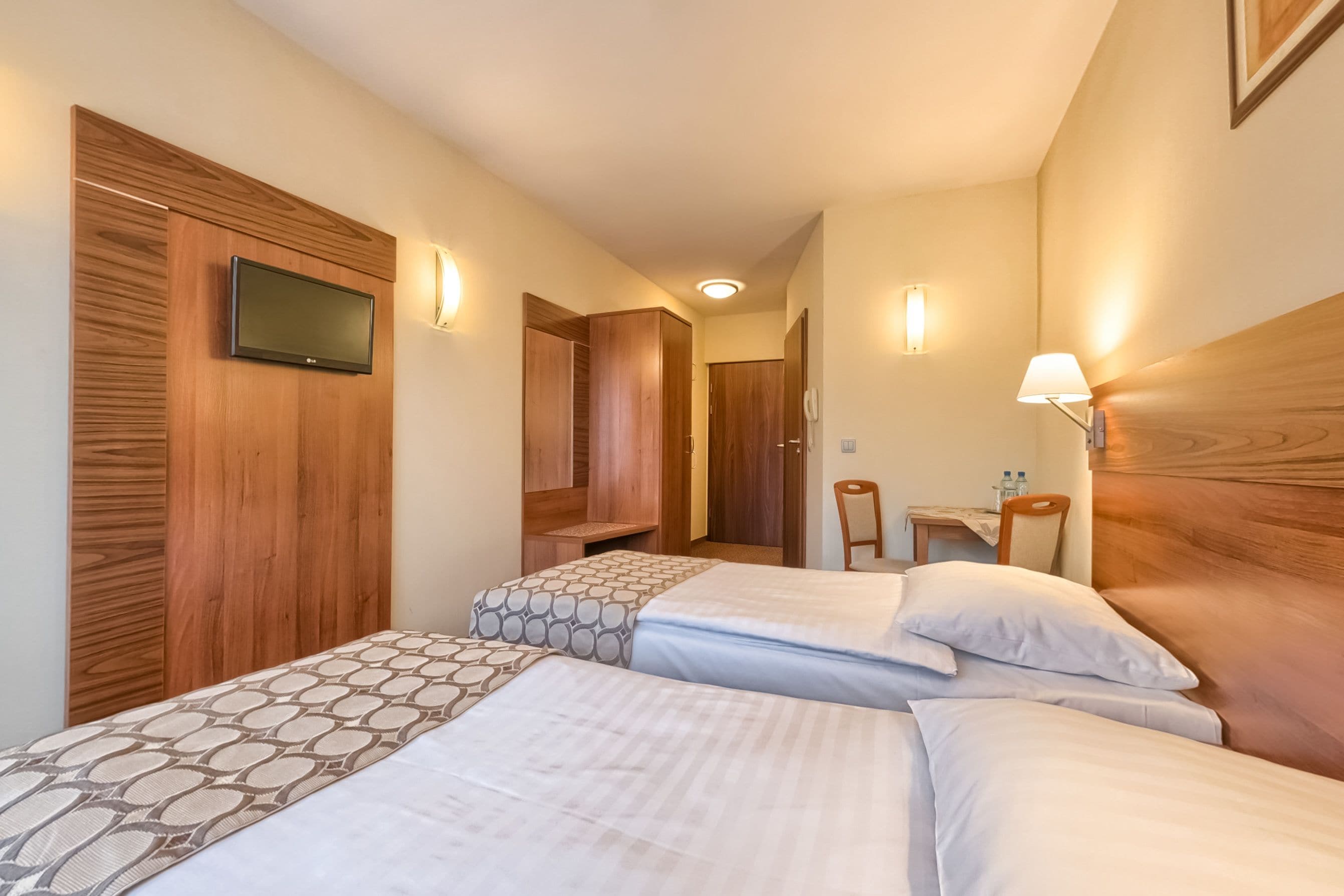 Een hotelkamer met twee bedden, een aan de muur bevestigde televisie en een houten garderobe.