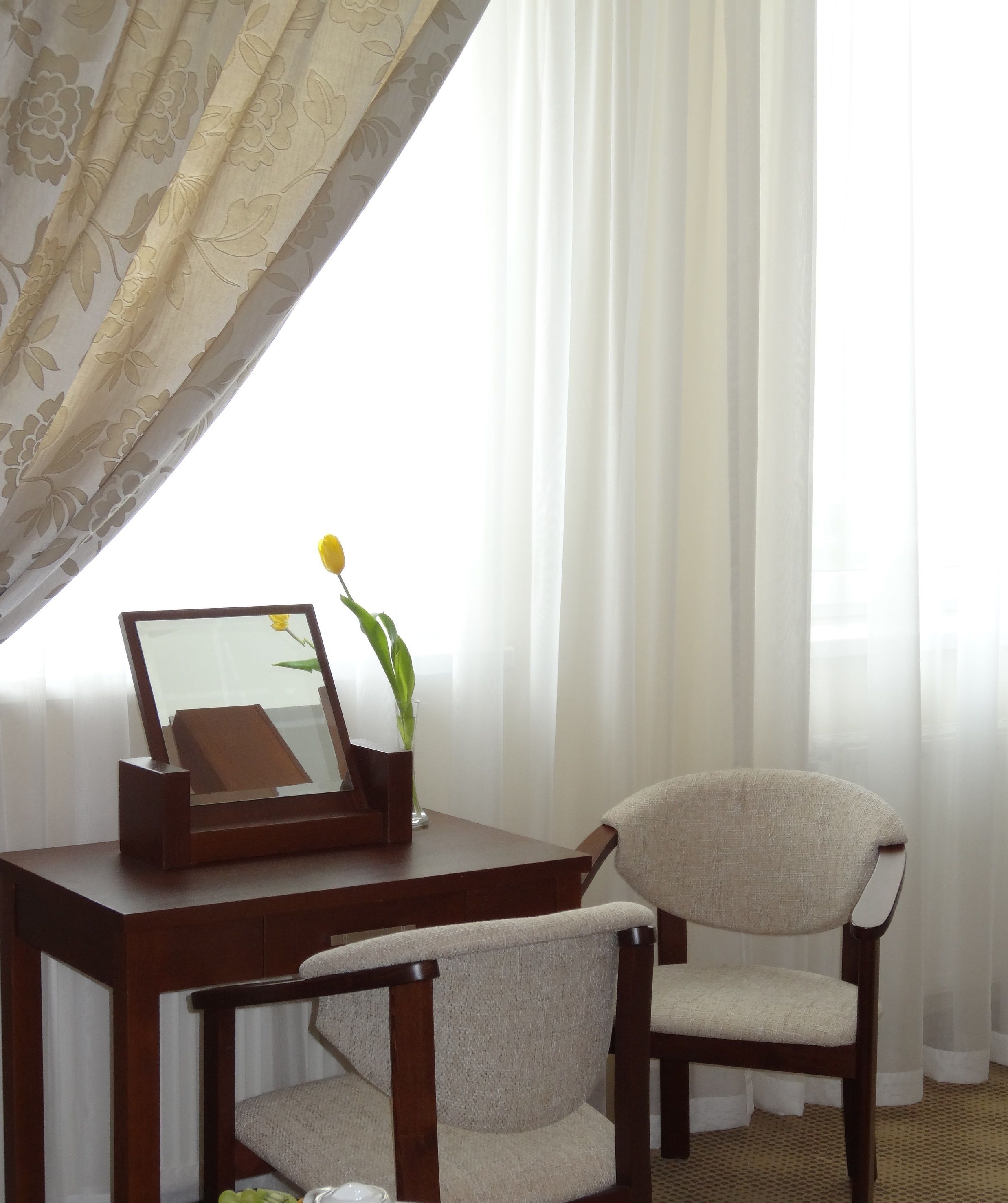 A room with a wooden desk, a chair, a mirror, and a vase with a yellow flower.
