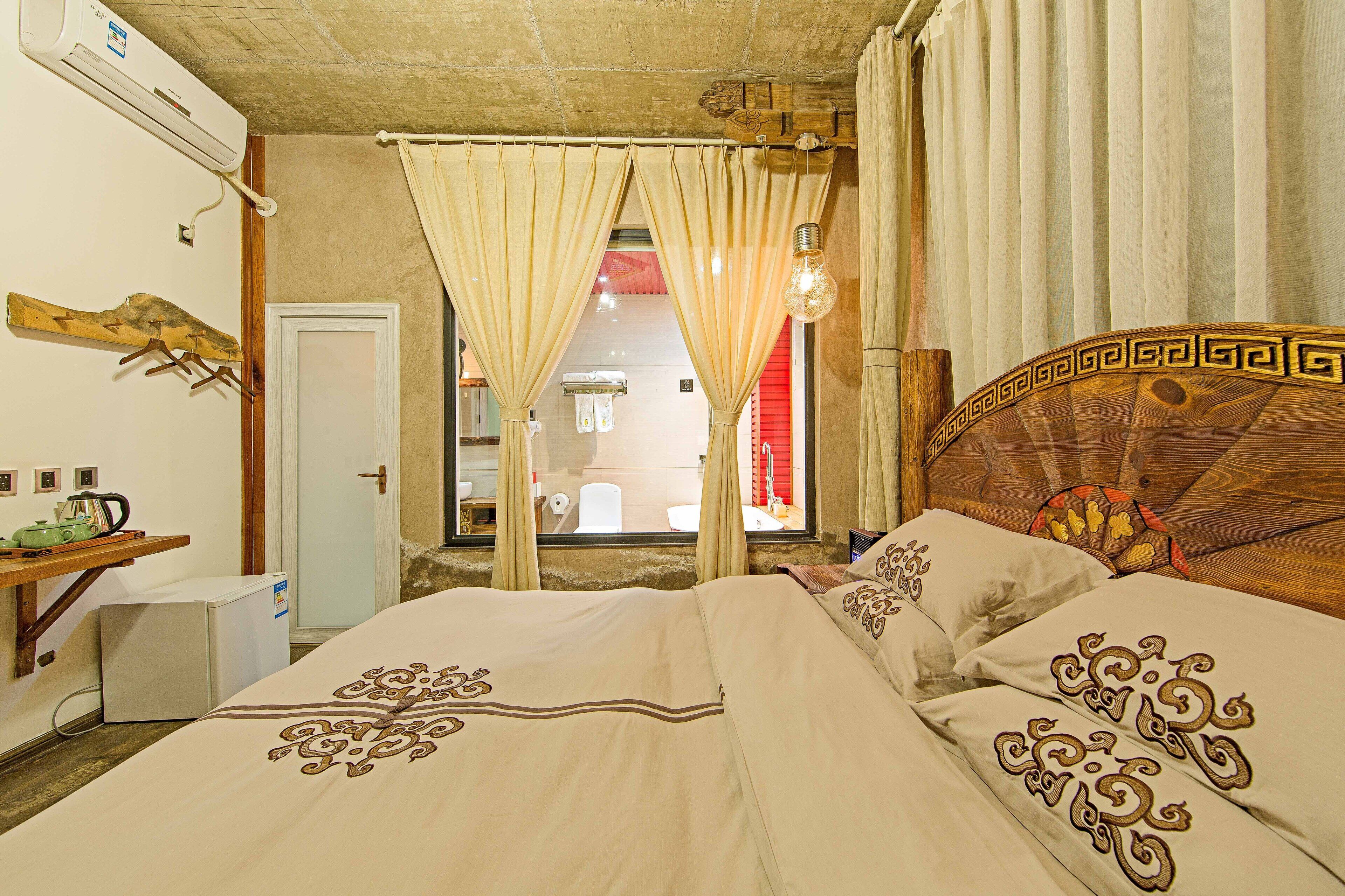 A bedroom with a bed, curtains, a wooden headboard, a small desk with a kettle, and a window with a view of the outside.