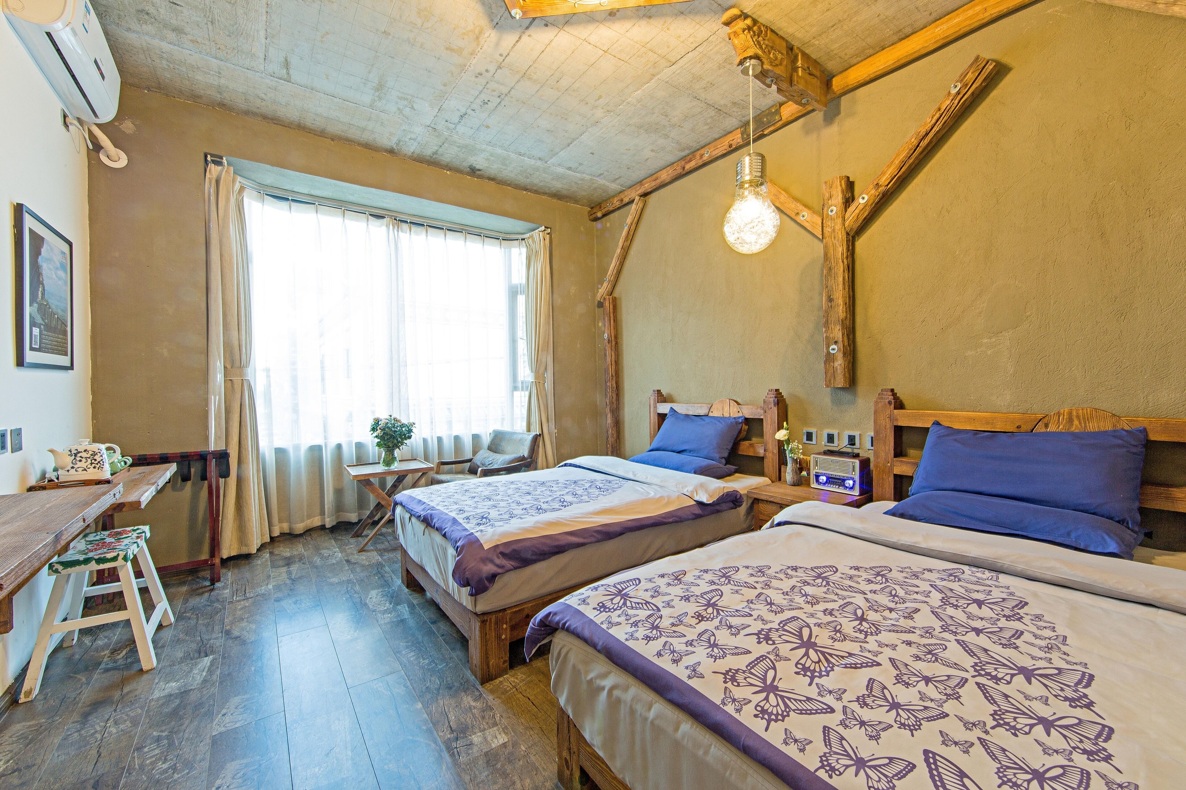 A room with two beds, a wooden desk, a chair, a window with curtains, and a hanging light fixture.