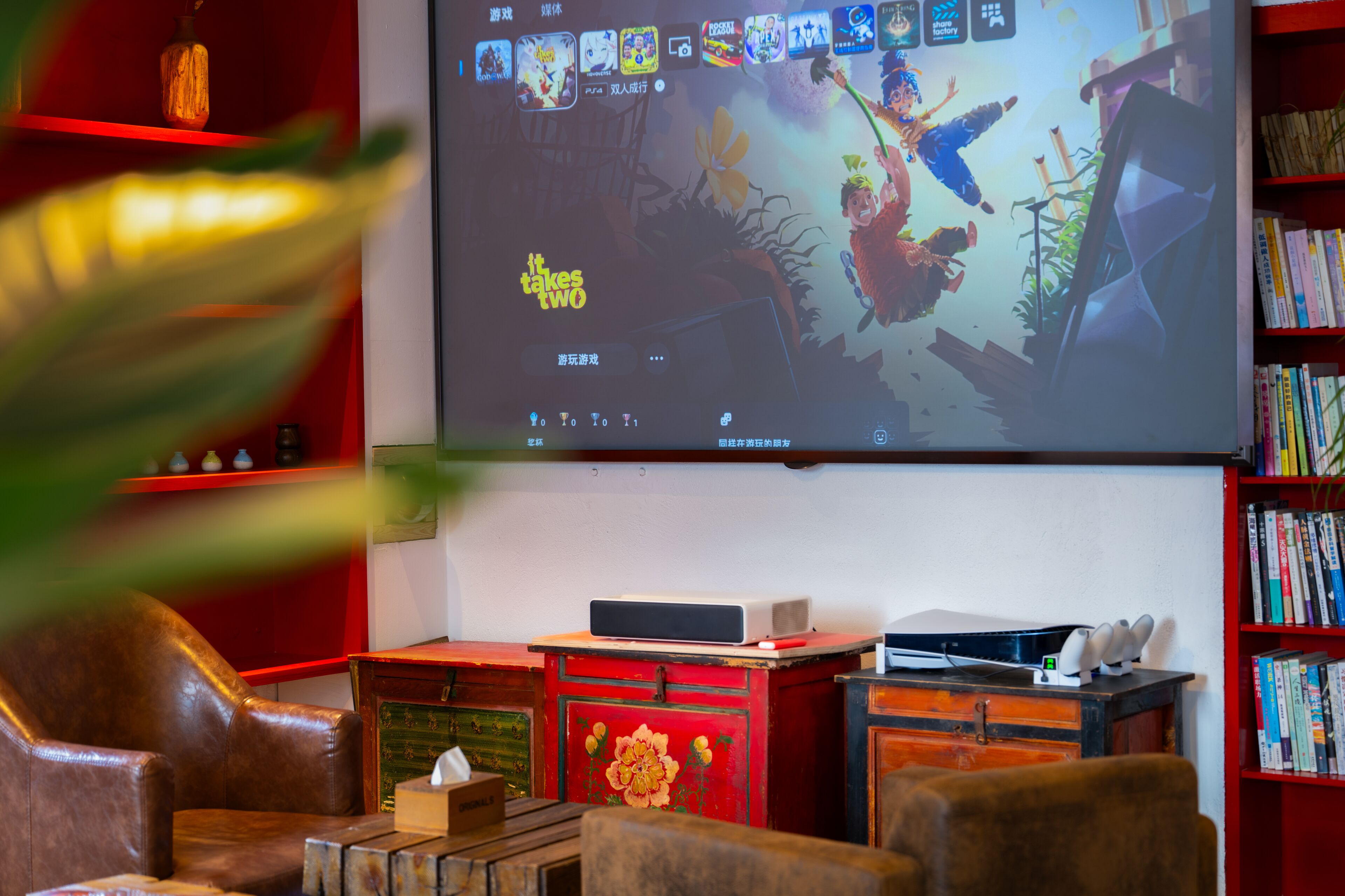 A room with a large screen displaying a game, a bookshelf, and a desk with a computer and speakers.