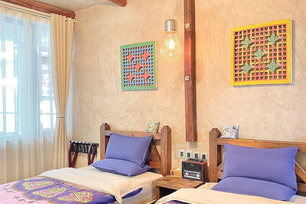 A room with two wooden beds, each with a purple pillow and a wooden headboard. There is a small table with a radio between the beds.