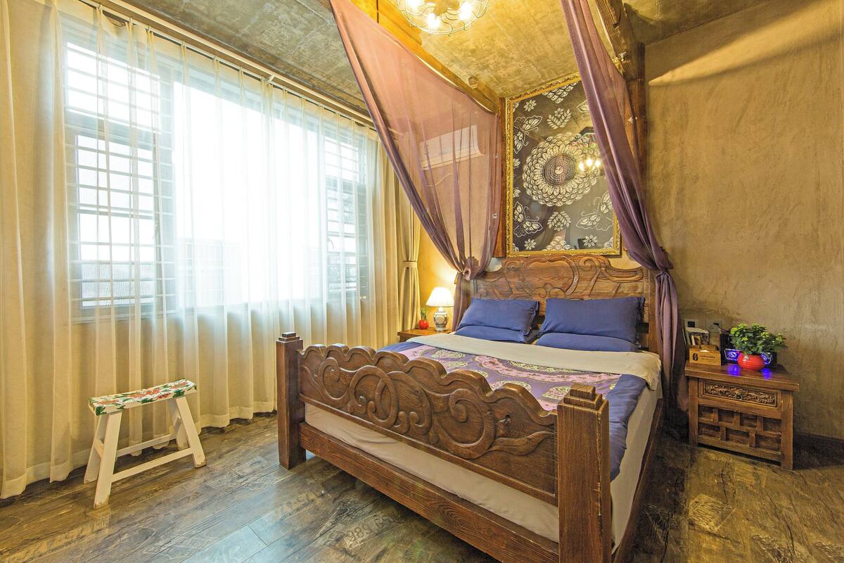 A wooden bed with a canopy, a small wooden stool, a nightstand with a lamp, and a decorative wall piece.