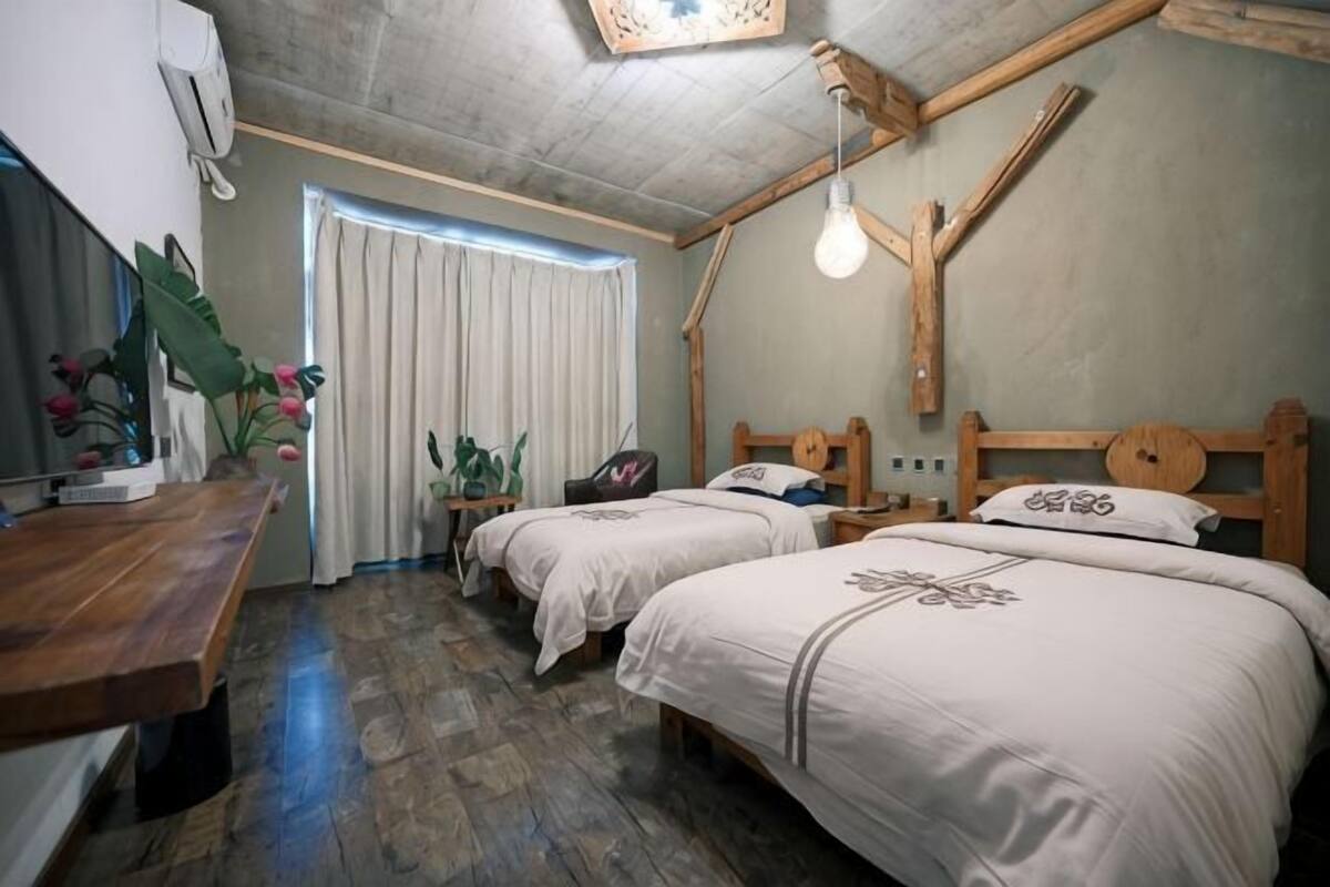 A room with two beds, a wooden desk, a window with curtains, and a ceiling light fixture.