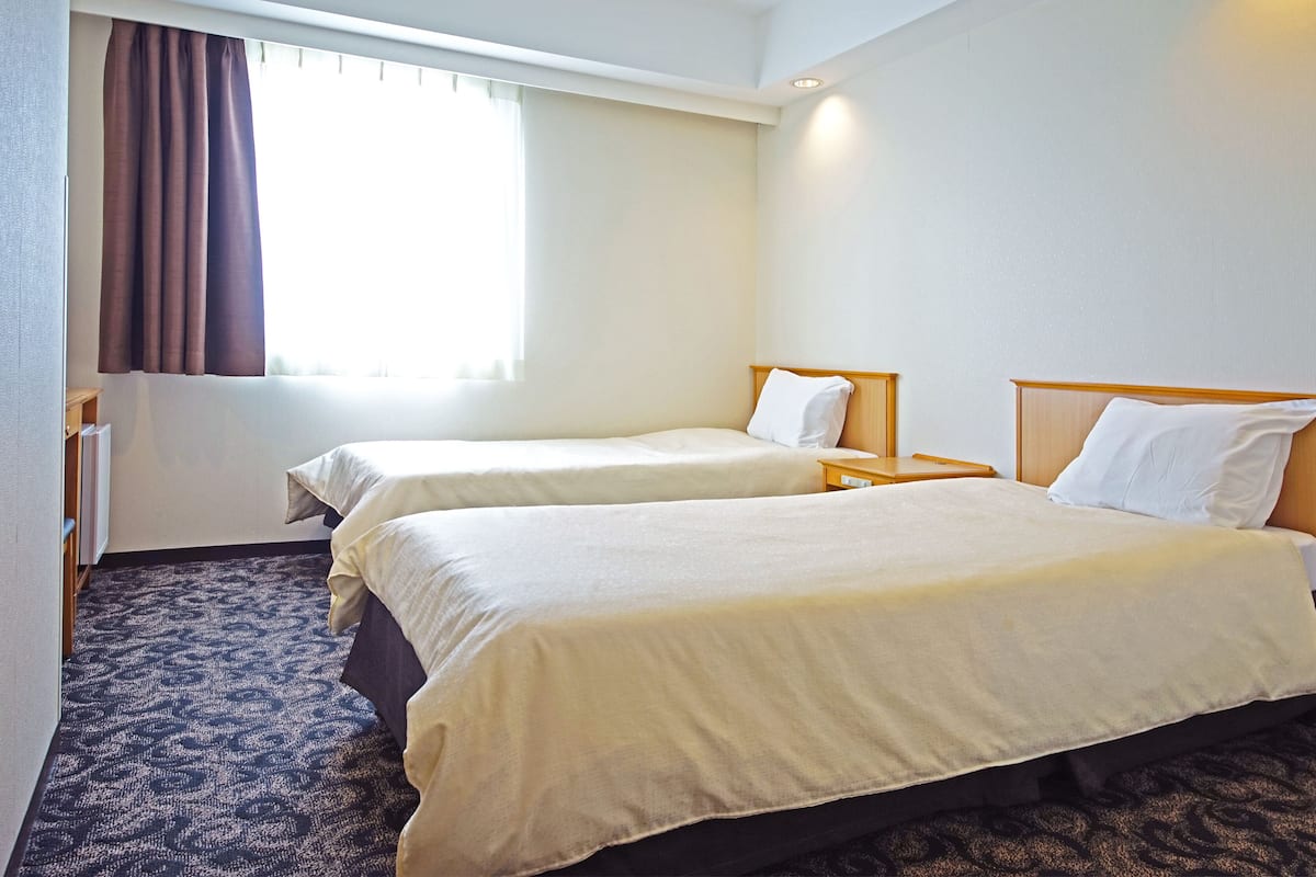 A hotel room with two single beds, a small bedside table, a window with curtains, and a carpeted floor.