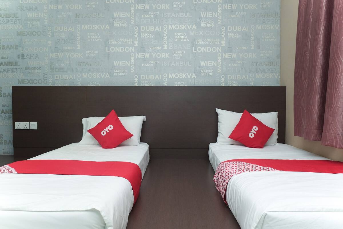 A hotel room with two beds, each with white bedding and red pillows, and a wall with city names in the background.