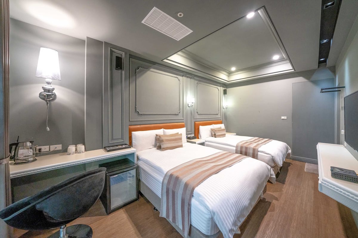 Una habitación de hotel con dos camas, un escritorio con lámpara, una silla y una kitchenette pequeña.