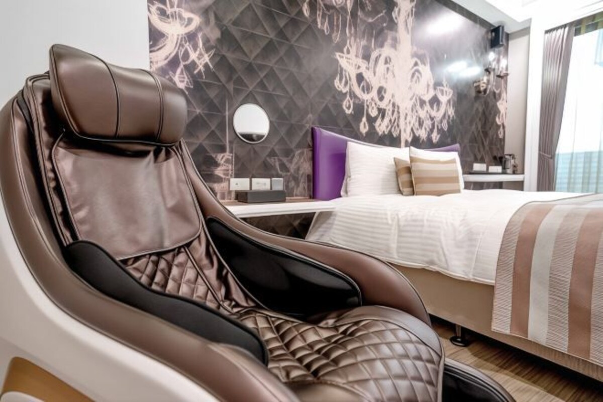 A modern hotel room with a massage chair, a bed with striped bedding, and a decorative wall with a geometric pattern.
