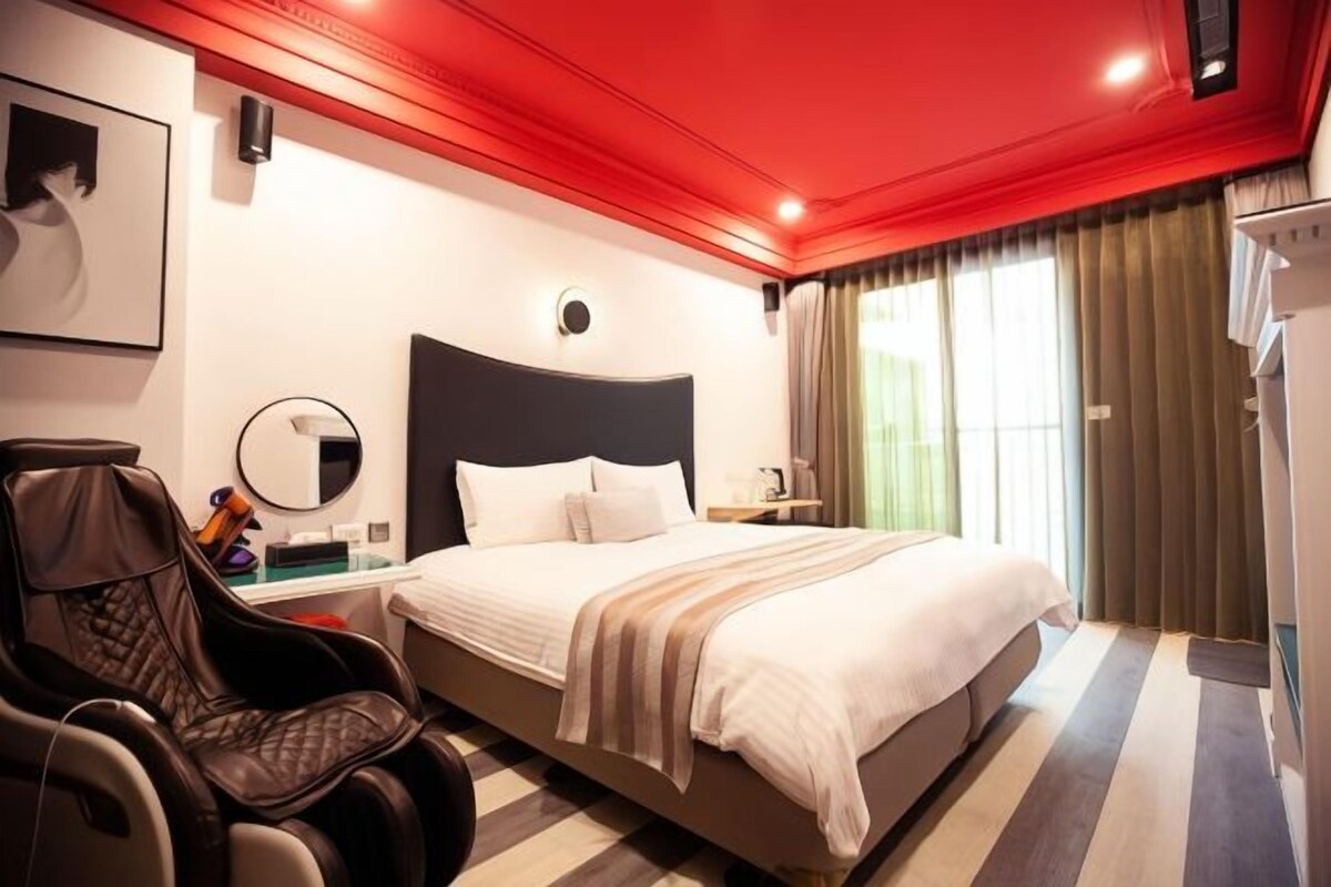 A hotel room with a large bed, a massage chair, a bedside table with a lamp, and a window with curtains.