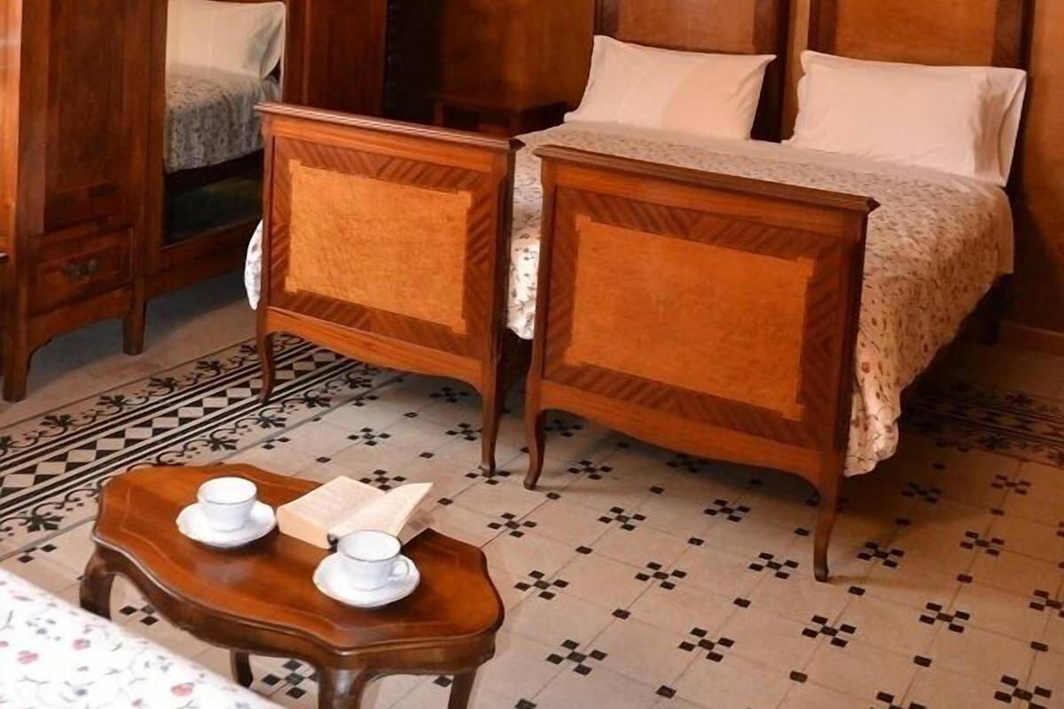 A room with two wooden beds, a wooden wardrobe, a small table with cups and a book, and a patterned floor.