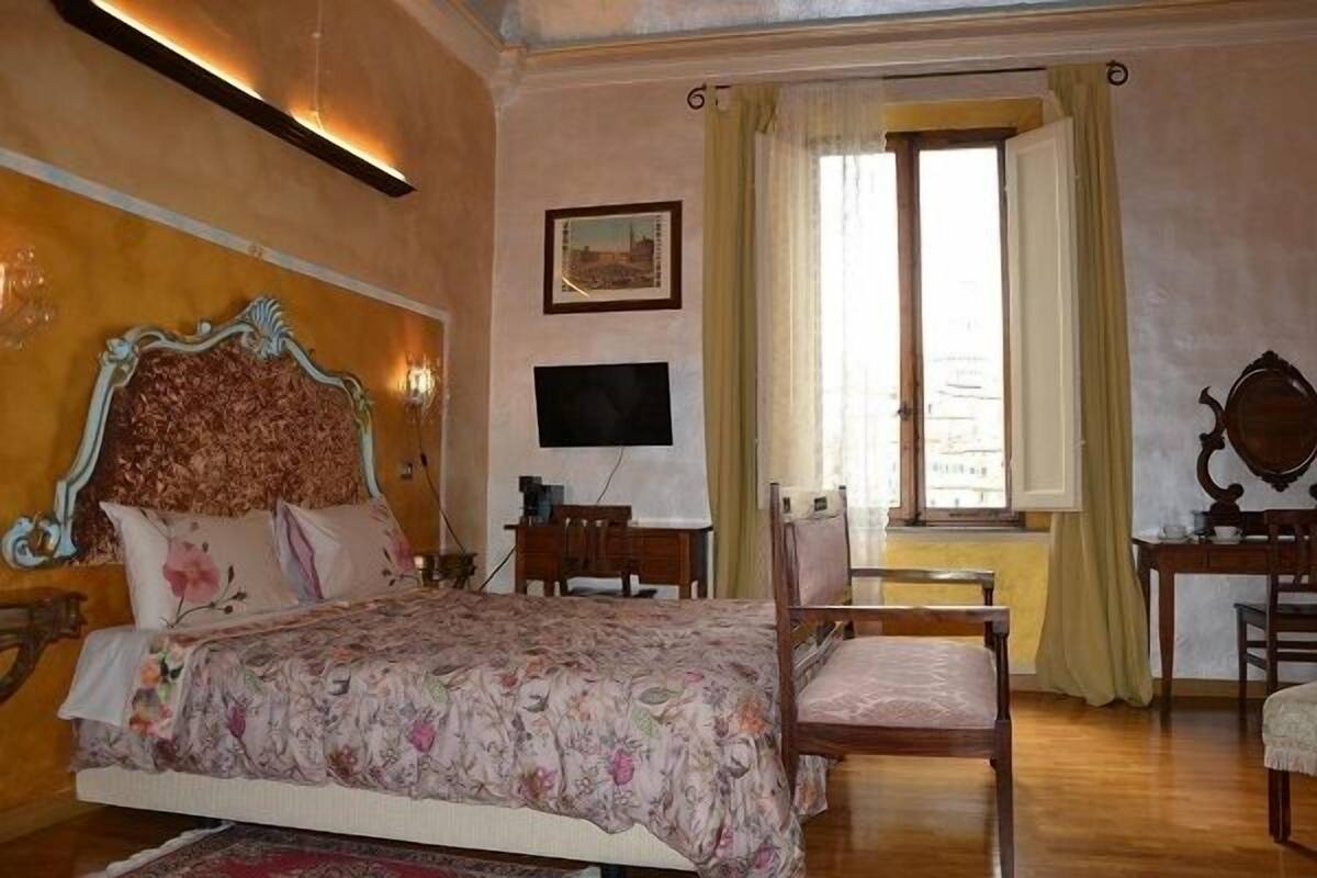 A bedroom with a large bed, a desk, a chair, a television, and a window with curtains.