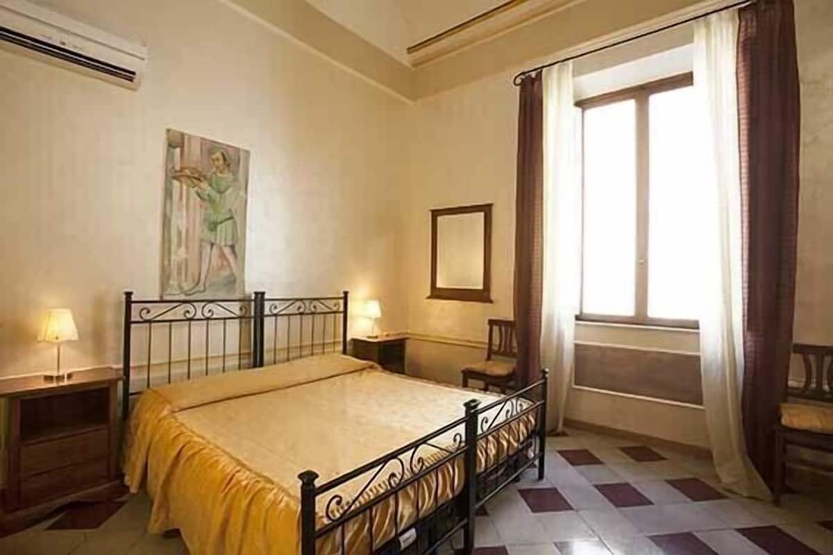 A bedroom with a bed, a nightstand, a lamp, a window with curtains, and a painting on the wall.