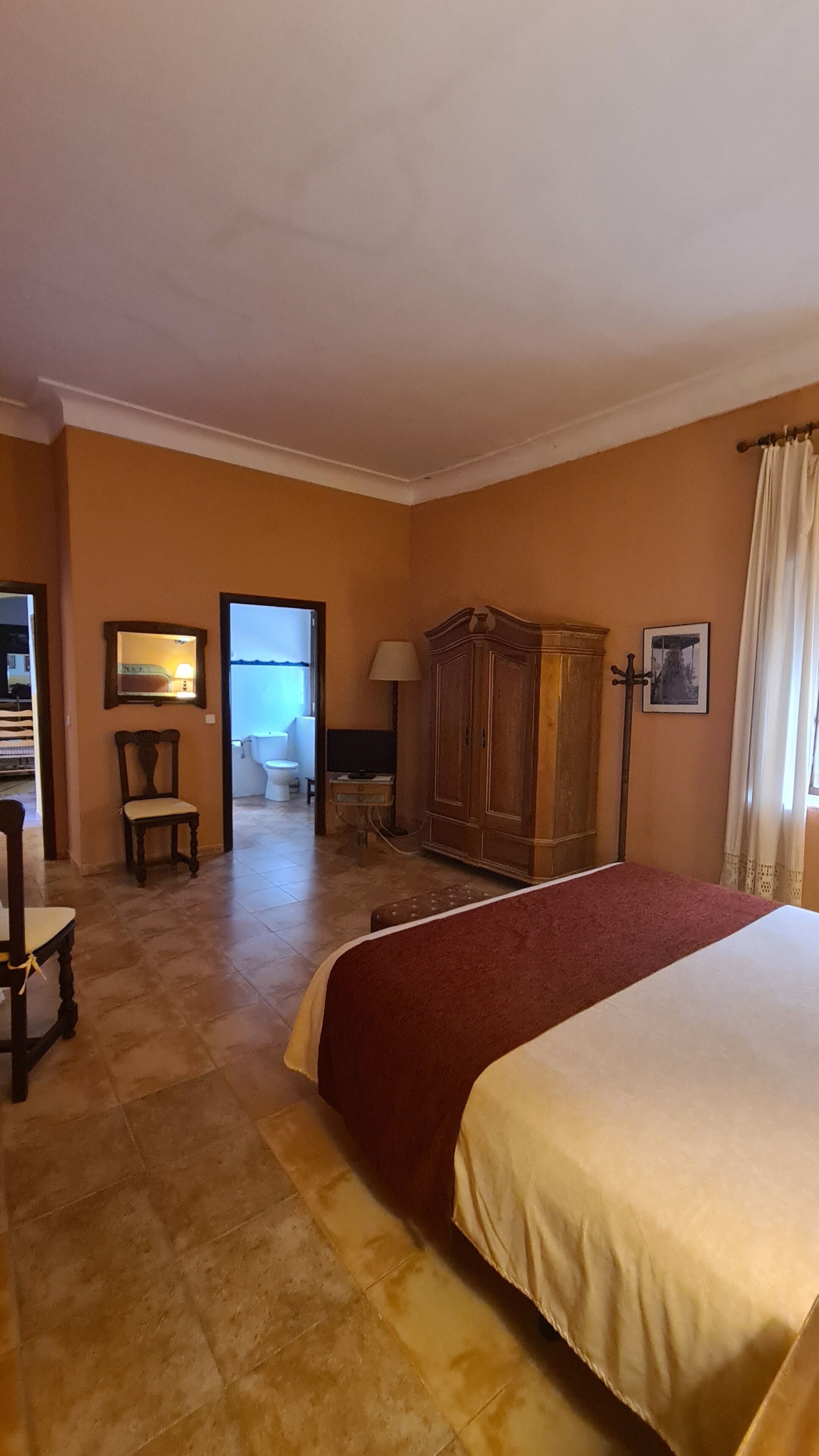 A hotel room with a large bed, a wooden wardrobe, a chair, a desk with a lamp, and a mirror on the wall.