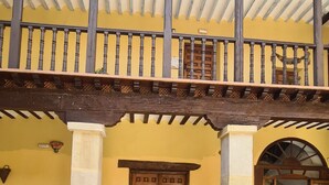 A building with a yellow facade, a wooden balcony with a railing, a double wooden door, and potted plants.