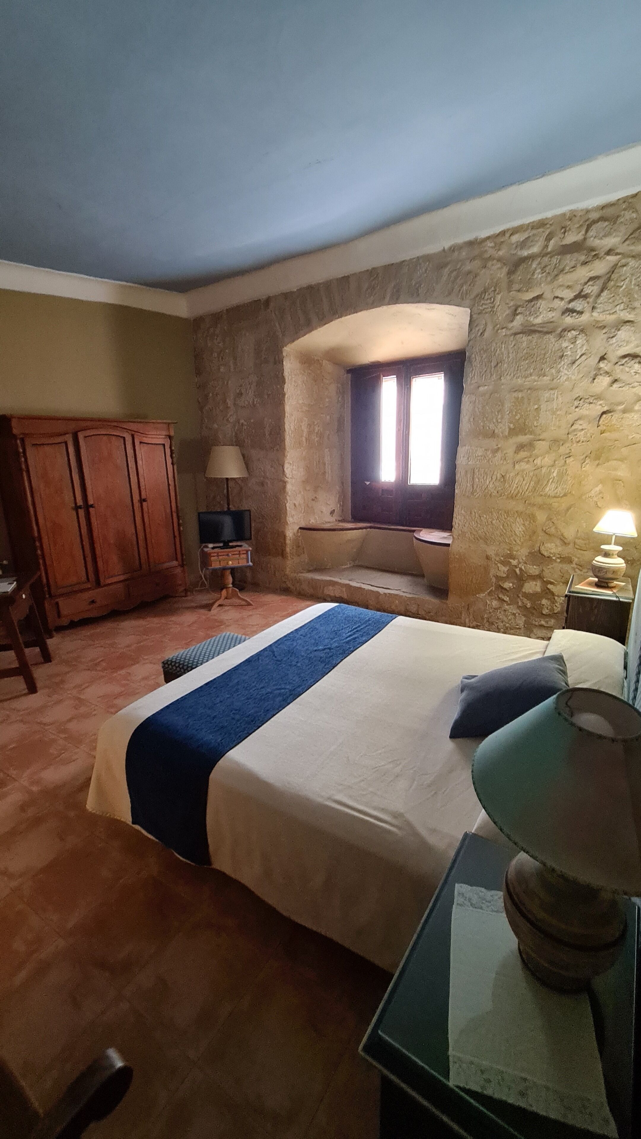 A bedroom with a bed, a wooden wardrobe, a stone wall, a window, and a lamp.