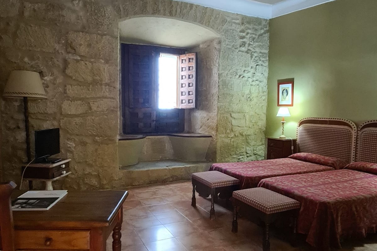 A room with a stone wall, a window with a view, a bed with a patterned bedspread, a wooden table, and a lamp.
