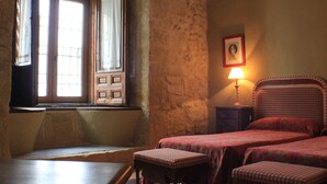 A room with a stone wall, a window with curtains, a bedside table with a lamp, and two beds with checkered bedding.