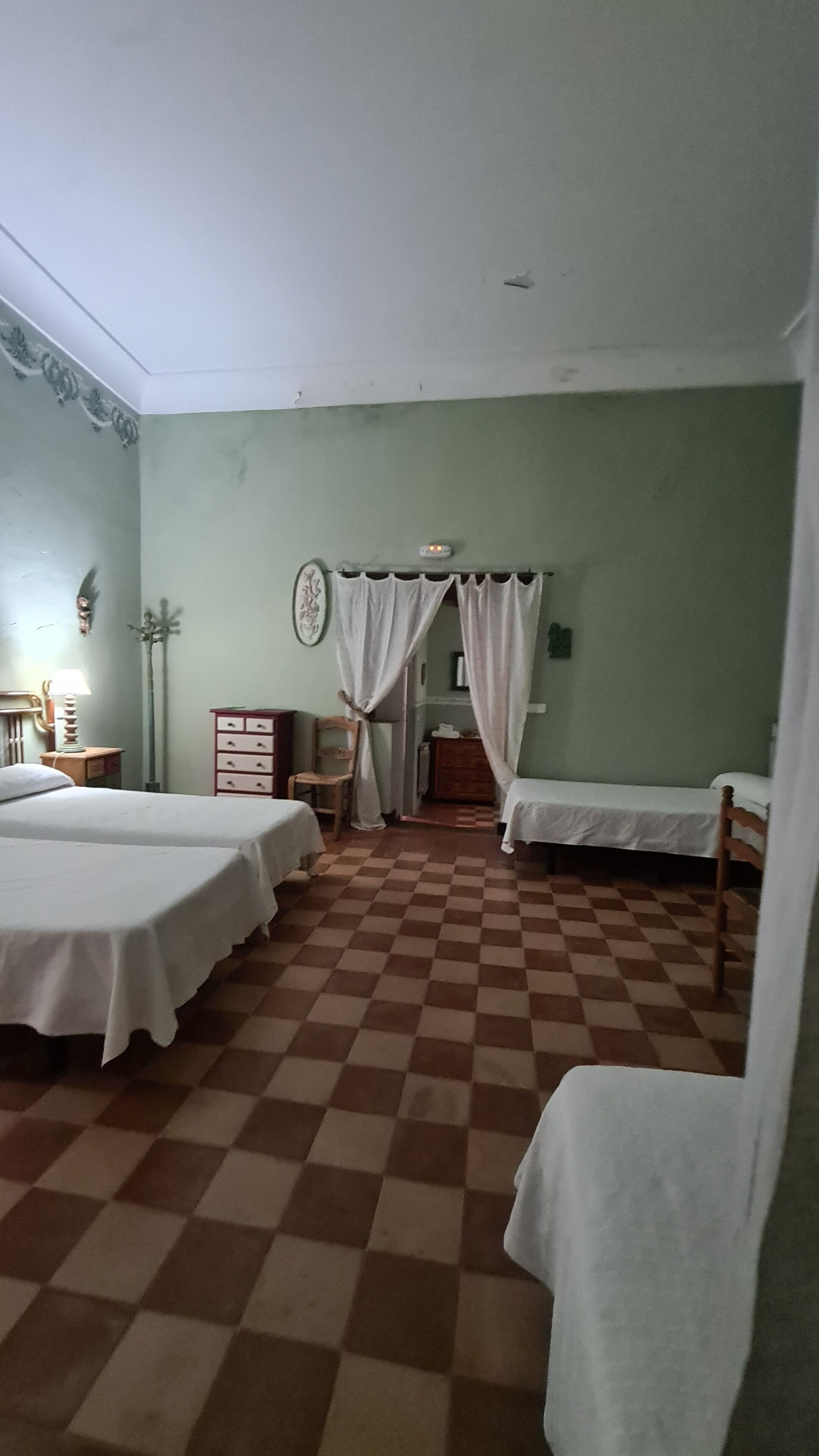 A room with two single beds, a wooden chair, a small table with a lamp, and a checkered floor.