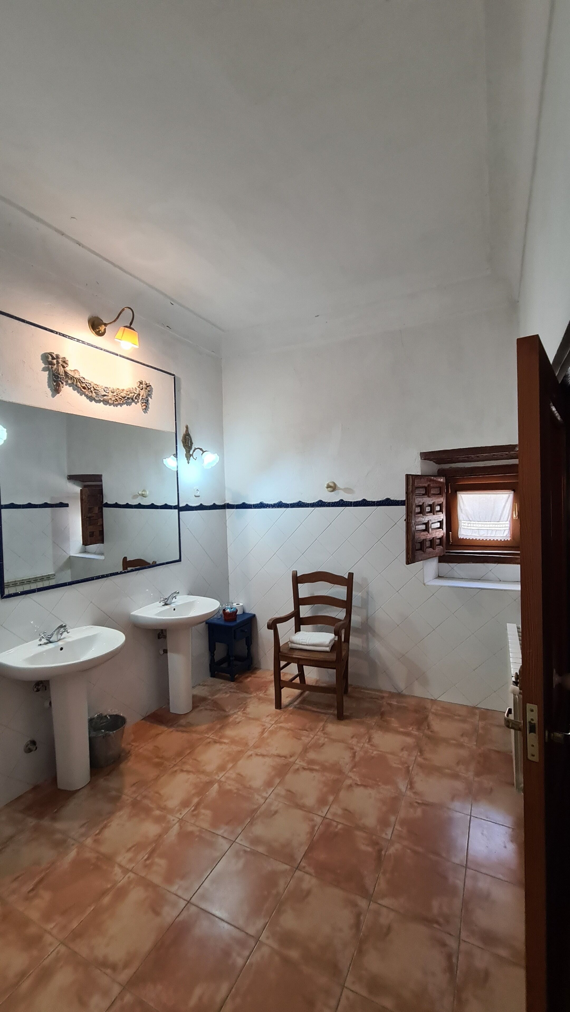 Un baño pequeño con dos lavamanos, un espejo, una silla de madera y una ventana con cortina.
