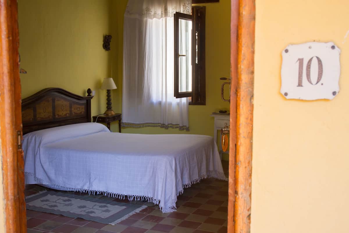 A bedroom with a bed, a nightstand, a lamp, a window with curtains, a wall-mounted number plate, and a tiled floor.