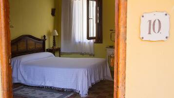 A bedroom with a bed, a nightstand, a lamp, a window with curtains, a wall-mounted number plate, and a tiled floor.