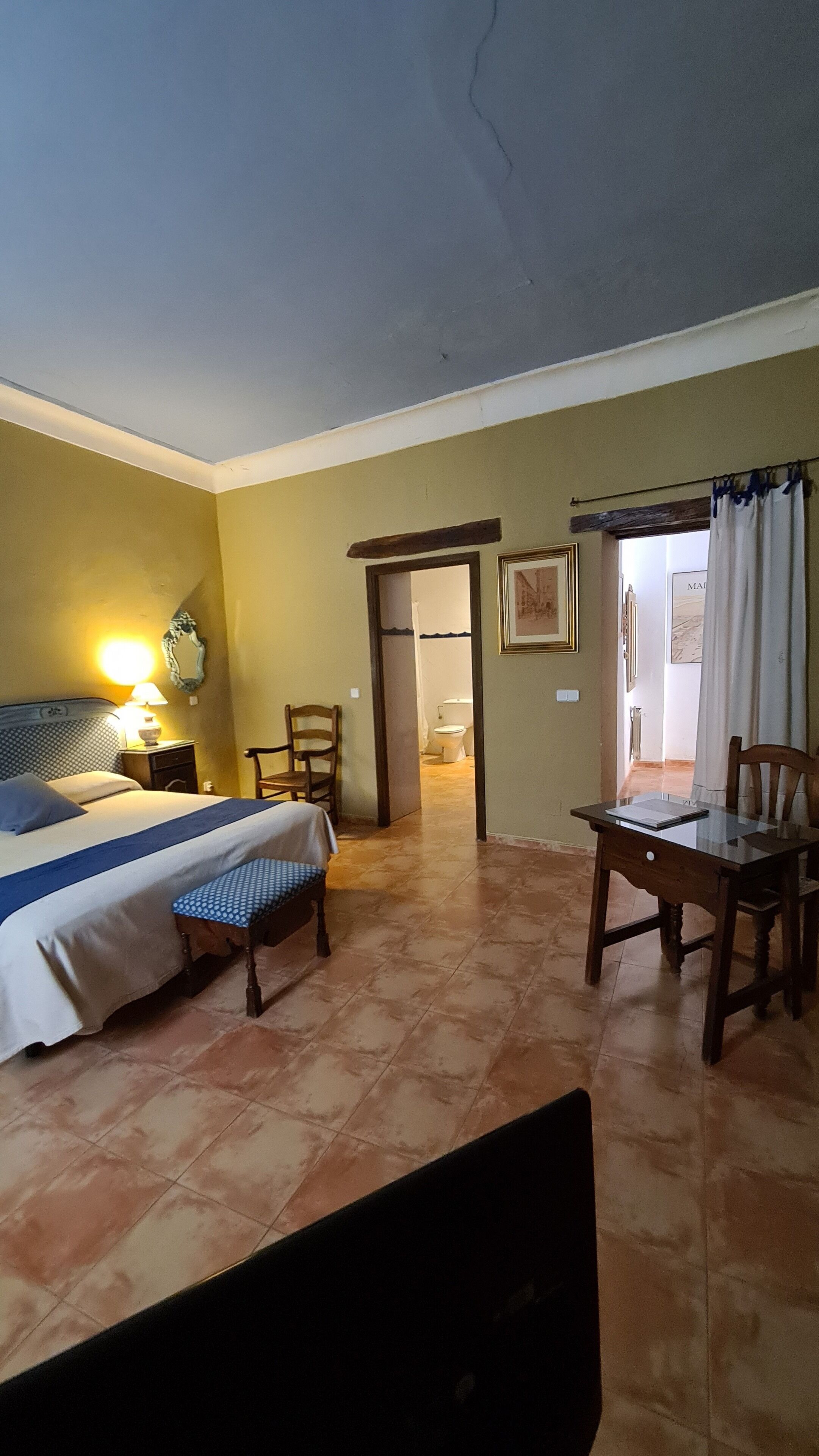 Habitación de hotel con cama, una mesita, una silla, un escritorio y un baño visible a través de una puerta abierta.