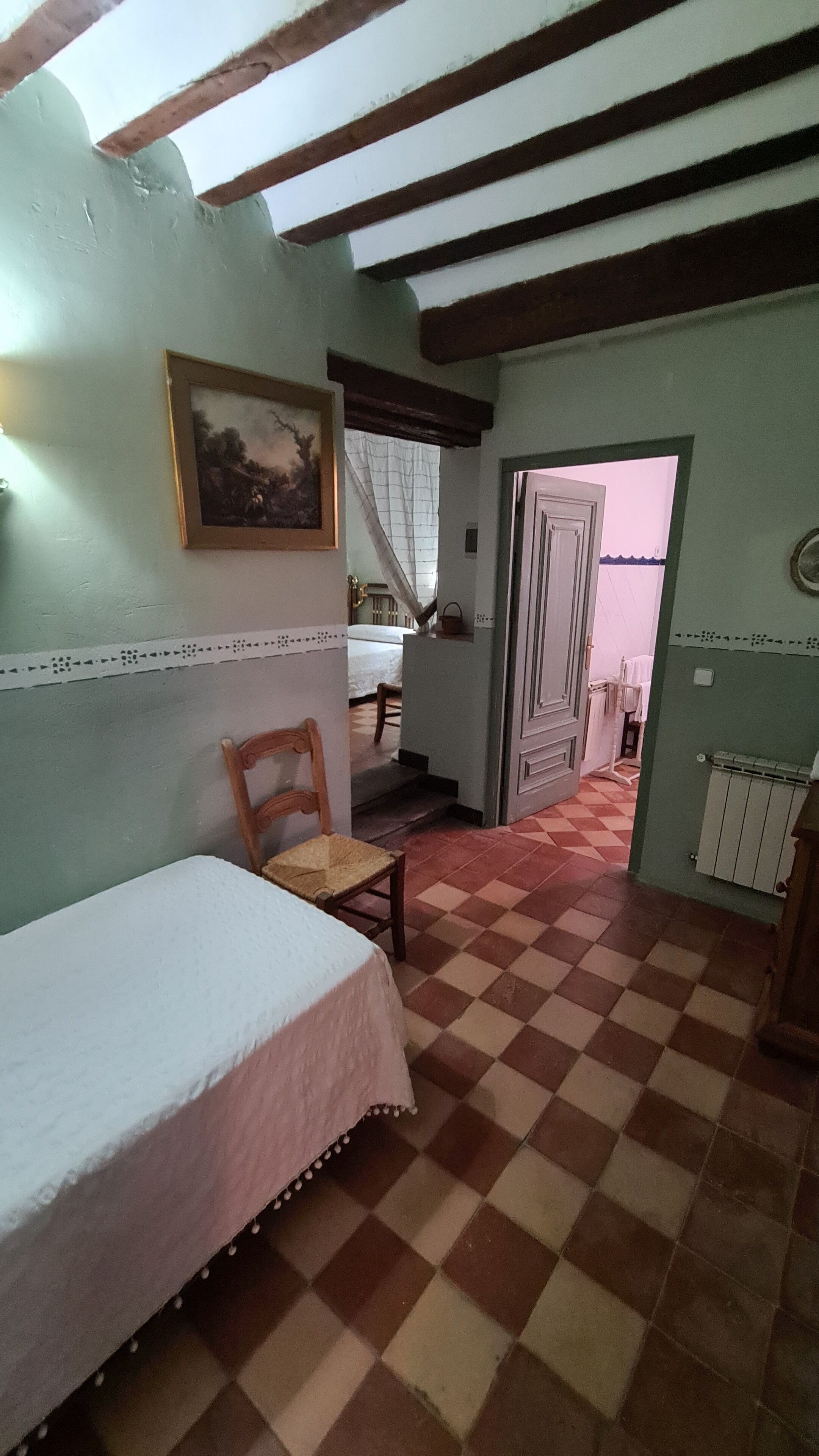 A room with a checkered floor, a bed, a chair, a painting on the wall, and a door leading to another room.