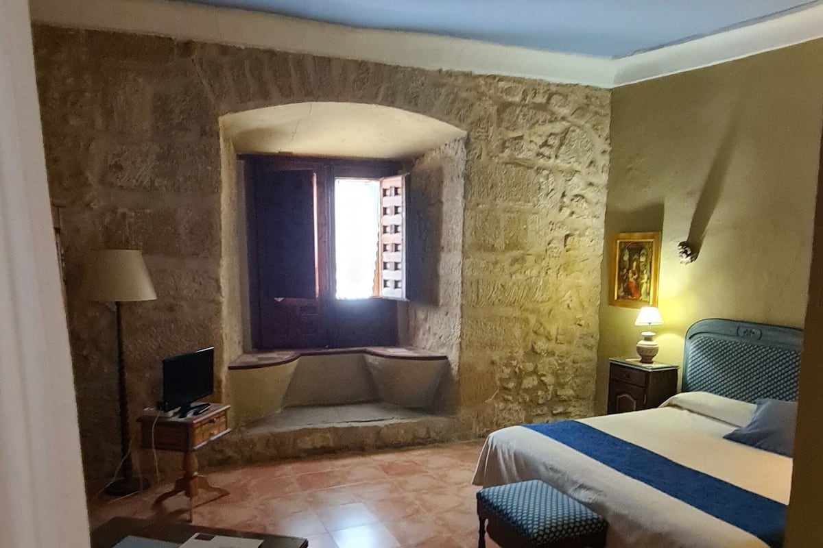 A bedroom with a stone wall, a bed, a nightstand with a lamp, a small table with a lamp, and a window with curtains.