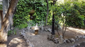 Un jardín con una mesa de piedra y sillas de metal, rodeado de follaje y un muro de piedra.