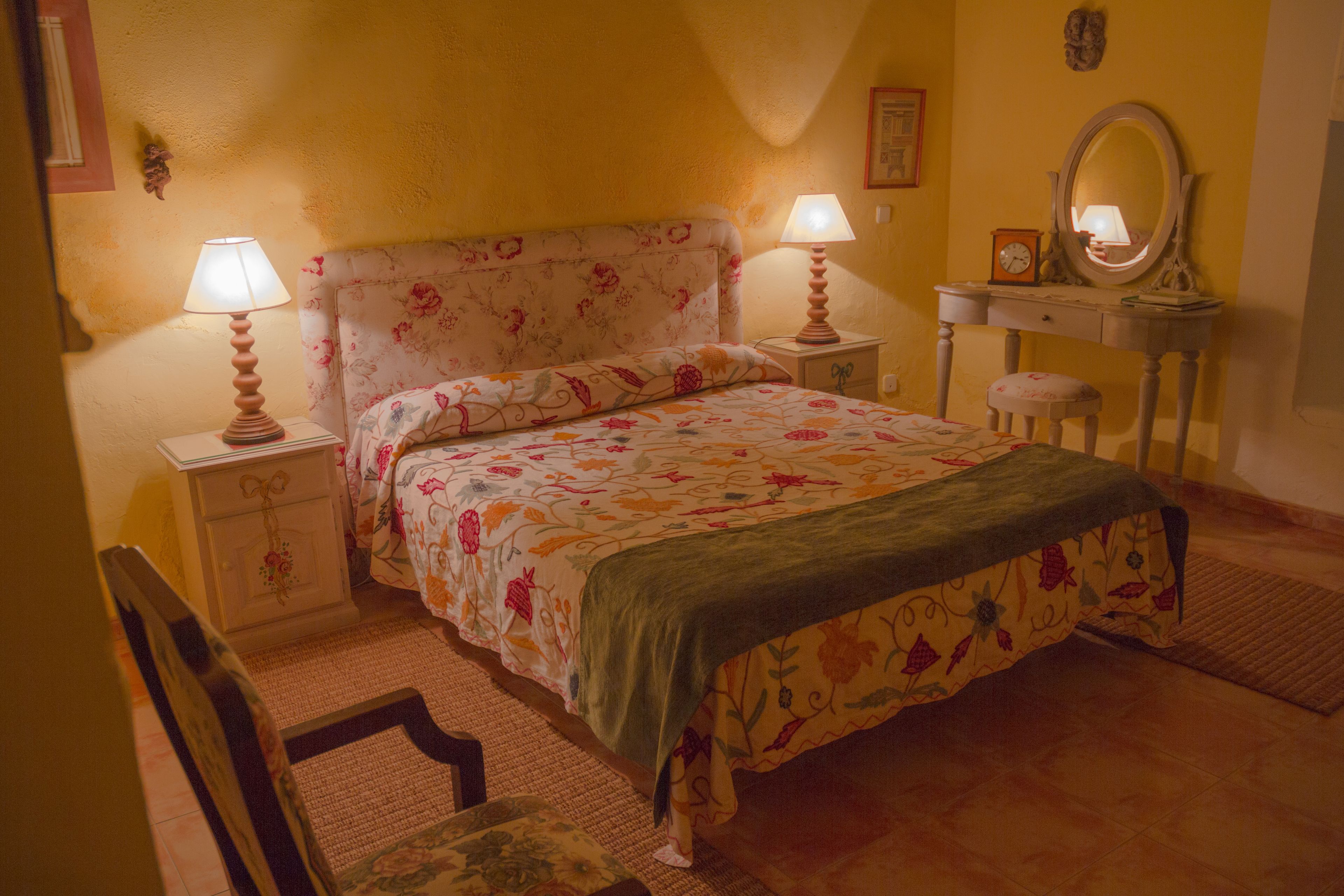 Un dormitori amb un llençol estampat de flors, dues làmpades de nit, un tocador amb mirall i una cadira.