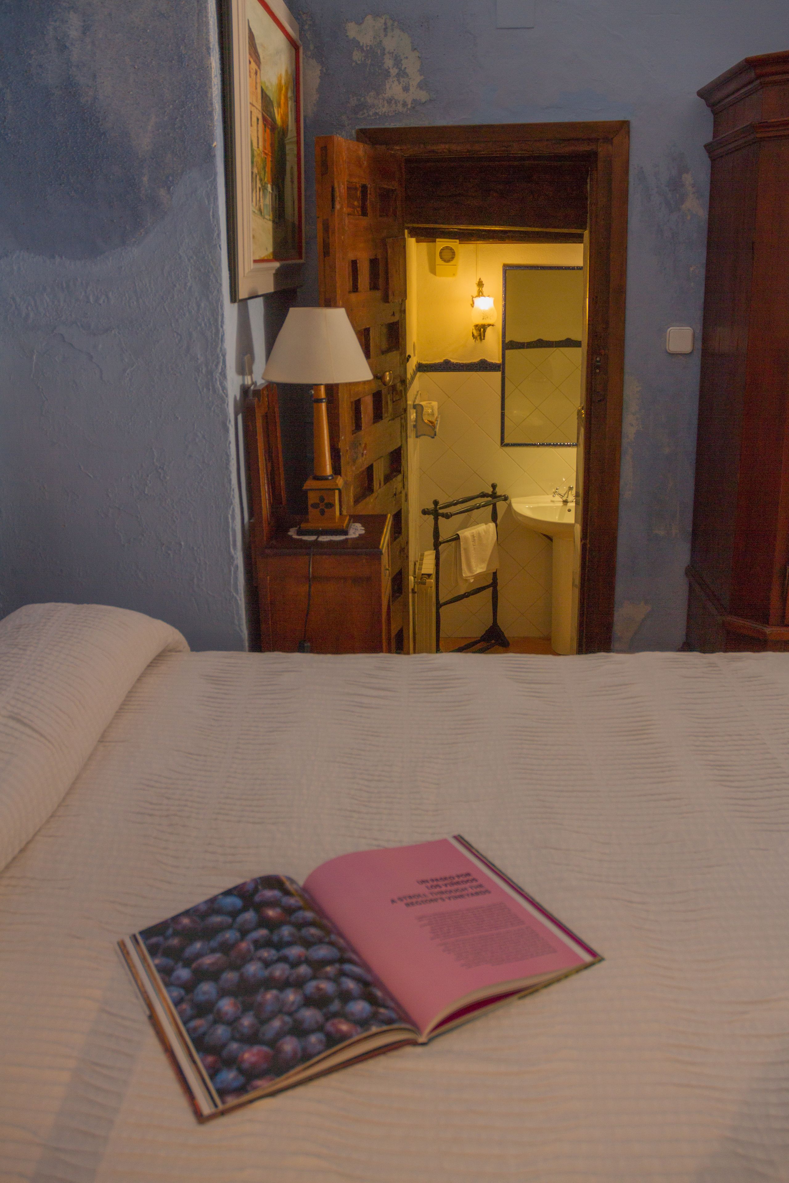 Un dormitorio con cama, una mesita de noche con lámpara, un libro y un baño visible a través de una puerta abierta.