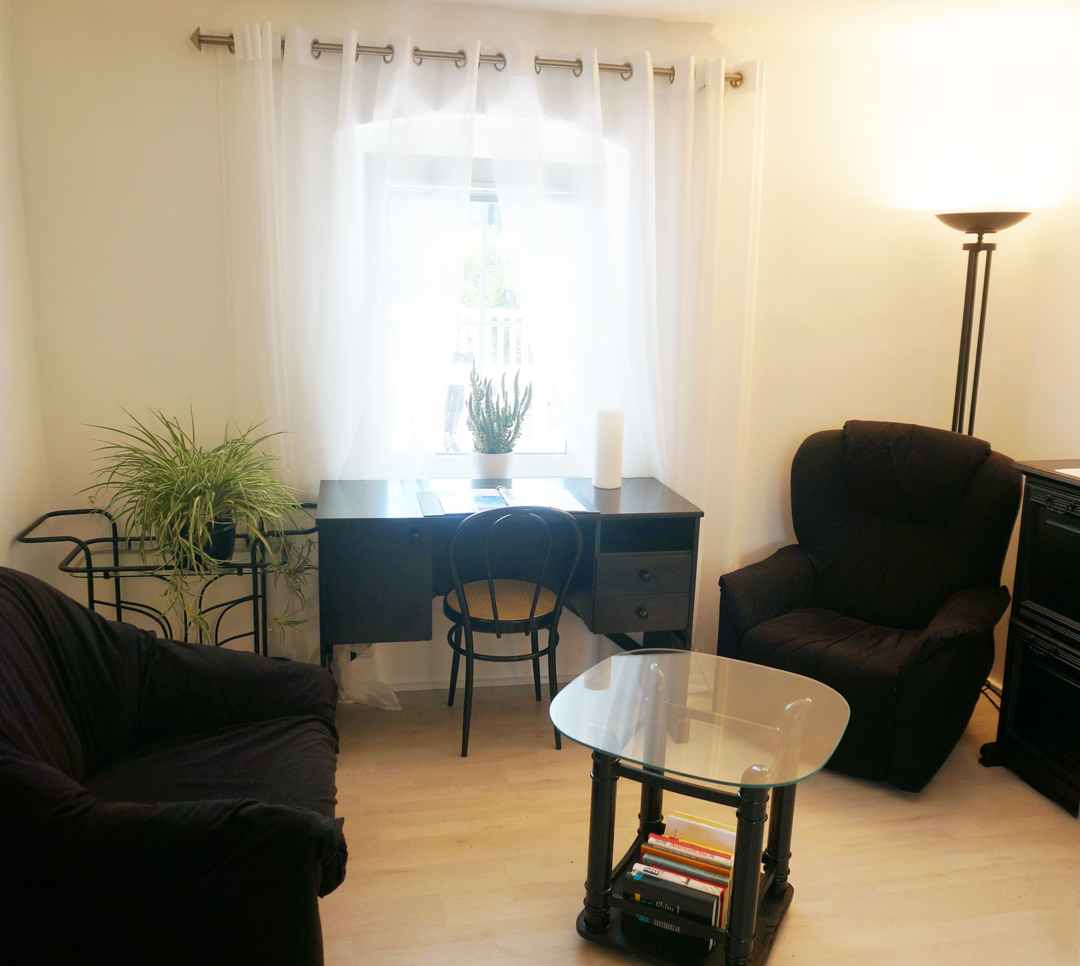 A small, well-lit room with a desk, a chair, a glass table with books, a potted plant, and a lamp.