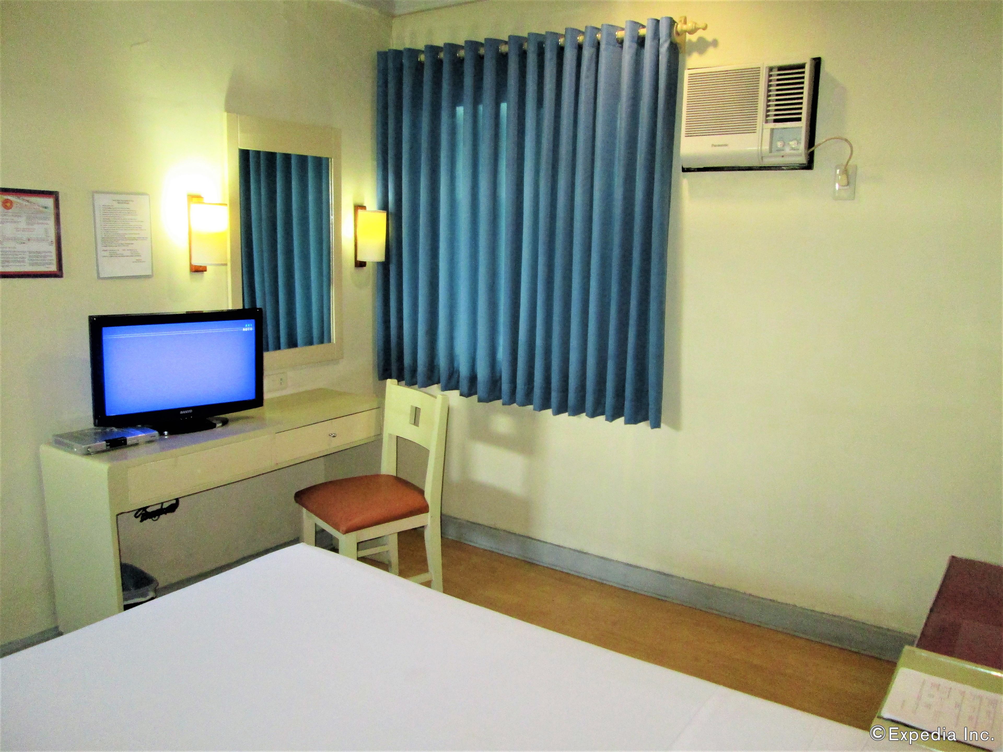 Une chambre d’hôtel comprenant un lit, un bureau avec un ordinateur, une chaise, une fenêtre avec des rideaux bleus et un appareil de climatisation.
