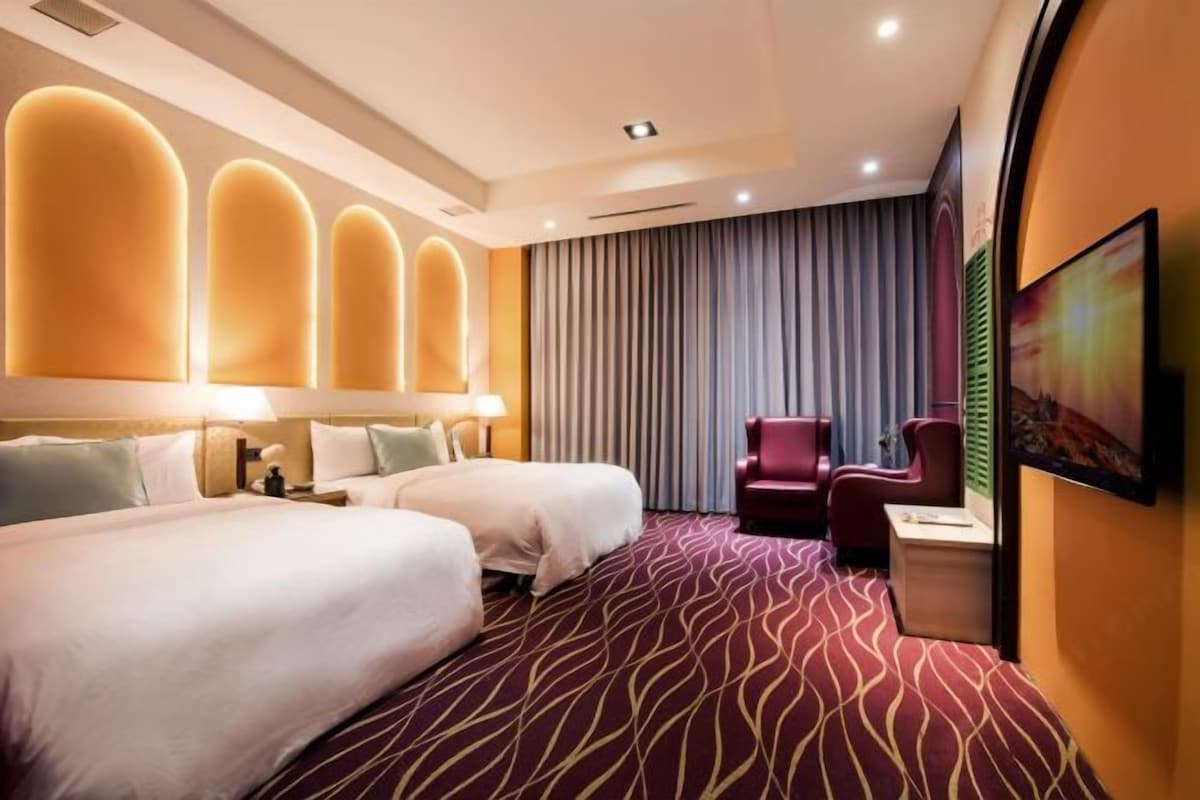 Une chambre d’hôtel avec deux lits, une moquette rouge, un fauteuil rouge et une grande fenêtre avec des rideaux.