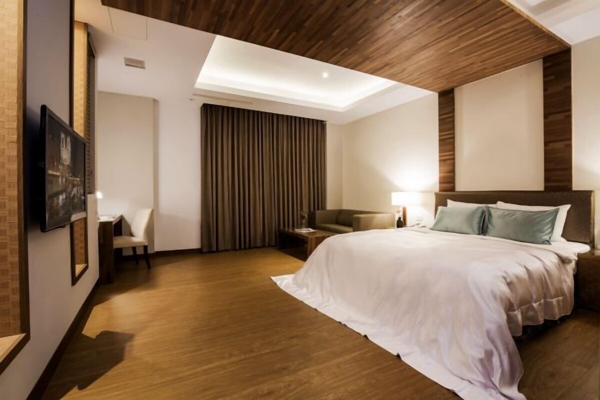 Une chambre d’hôtel moderne avec un plafond en bois, un grand lit avec une literie blanche, un coin salon avec une chaise et une table, et une grande fenêtre avec des rideaux.