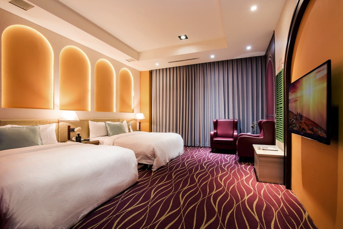 Een hotelkamer met twee bedden, een rode fauteuil, een bureau en een groot raam met gordijnen.