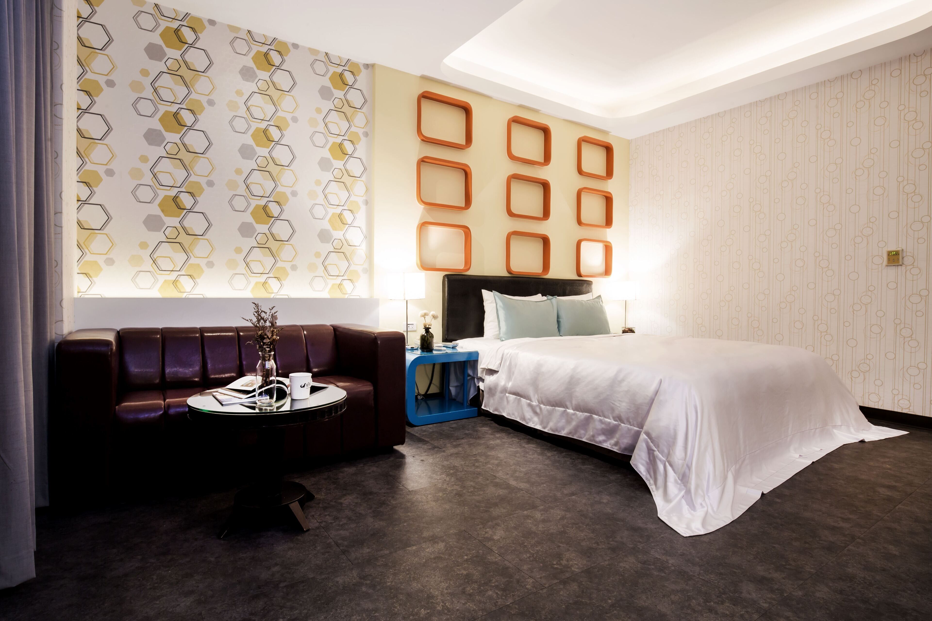 Une chambre d’hôtel moderne avec un canapé en cuir marron foncé, un lit blanc avec une couette blanche, une table de chevet bleue et un mur orné de motifs géométriques.