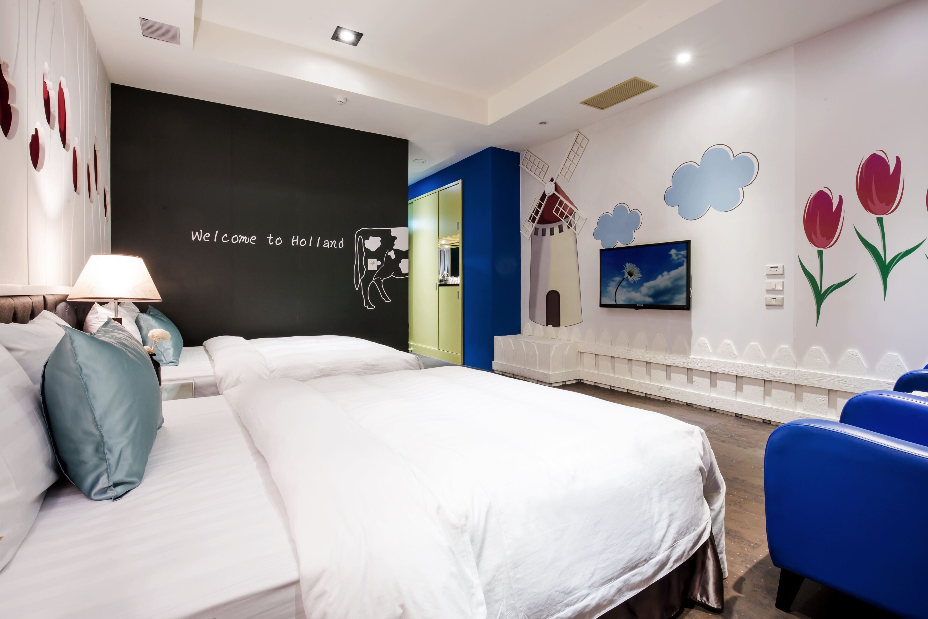 A hotel room with a bed, a desk, a chair, a lamp, and a wall with a welcome sign and a windmill painting.