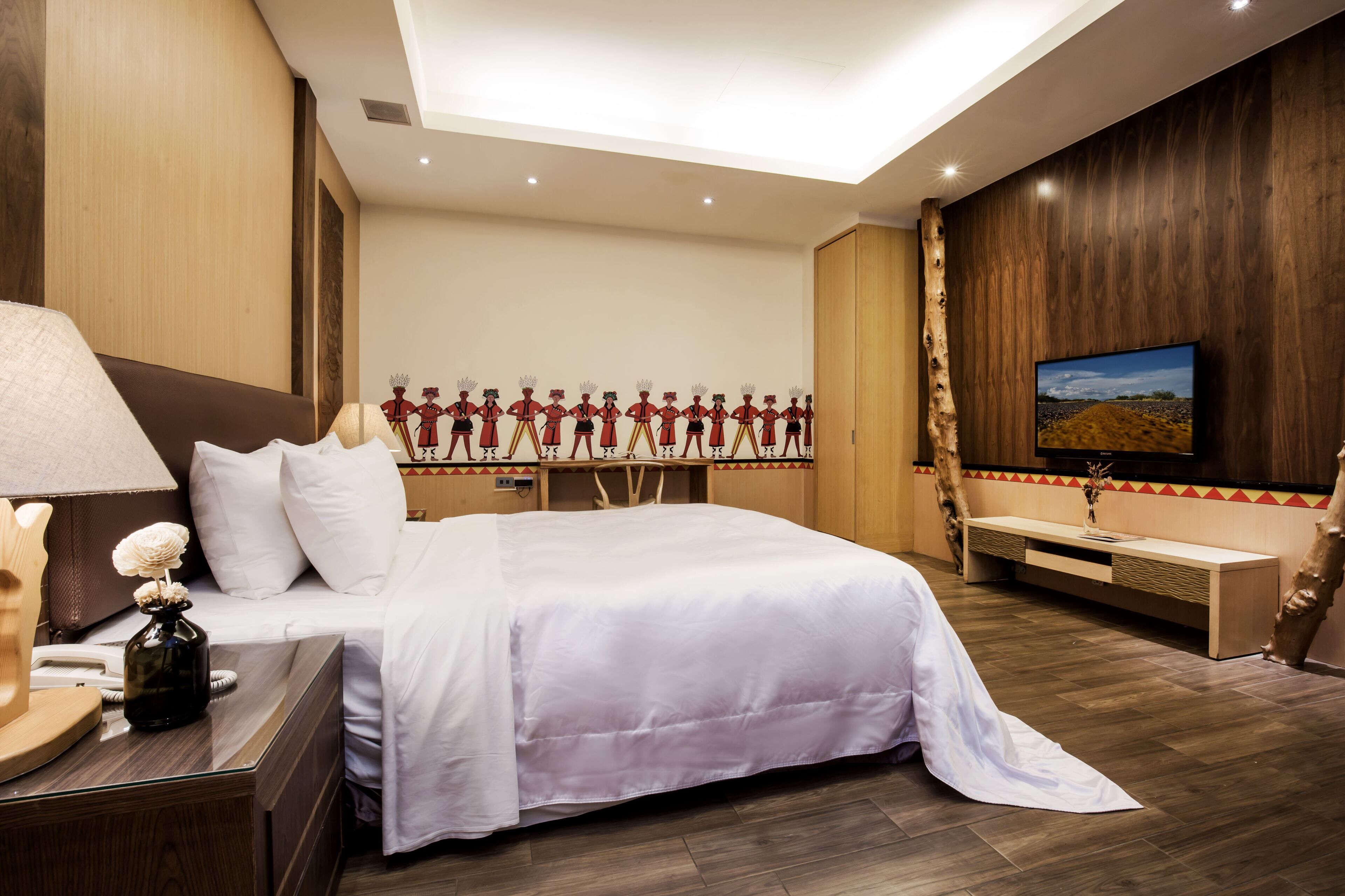 Una habitació d'hotel amb un llit gran, un panell de fusta al capçal, una taula de nit amb una làmpada, un televisor fixat a la paret i un panell decoratiu amb figures.