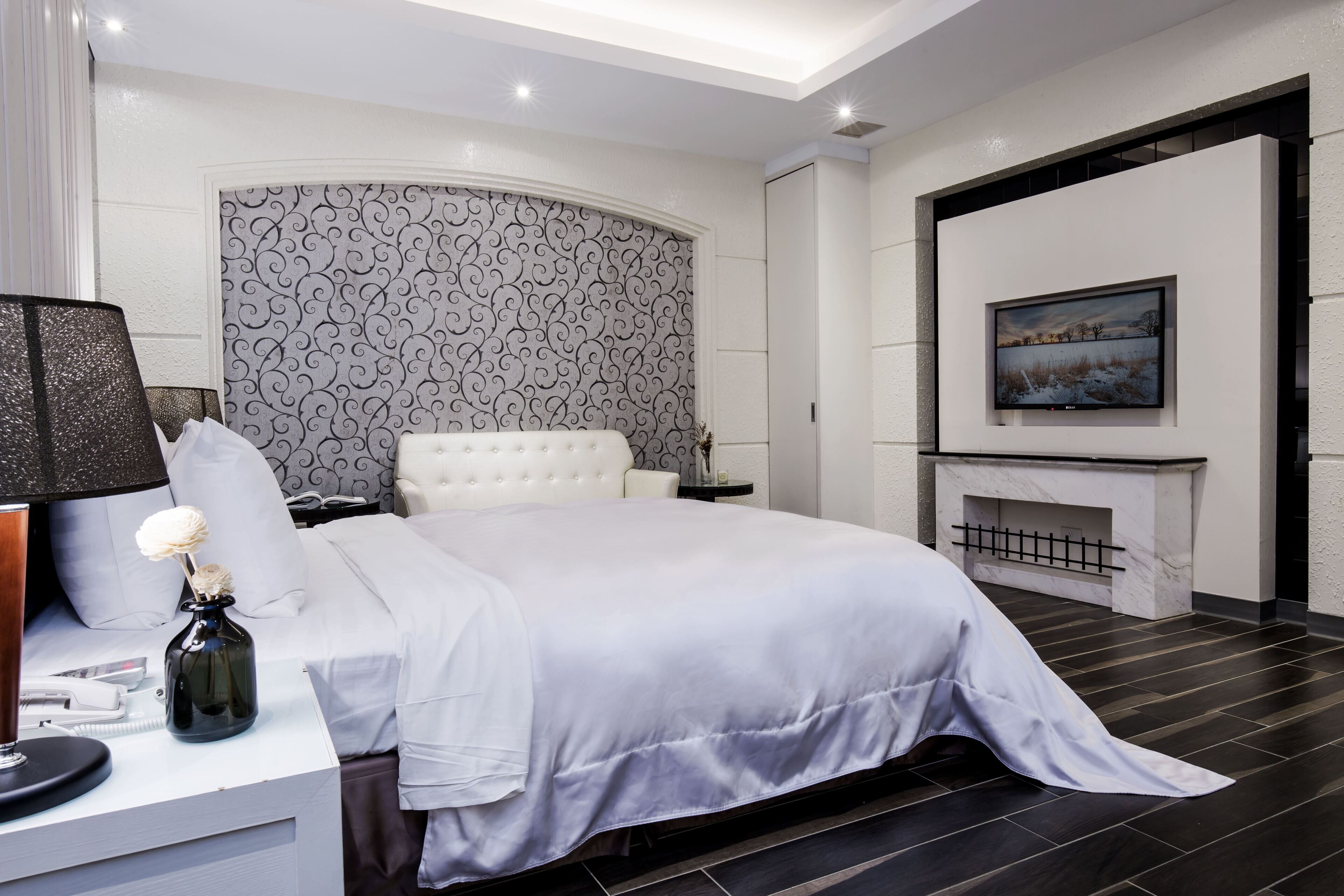 A modern bedroom with a large bed, a bedside table with a lamp, a decorative vase, and a fireplace.