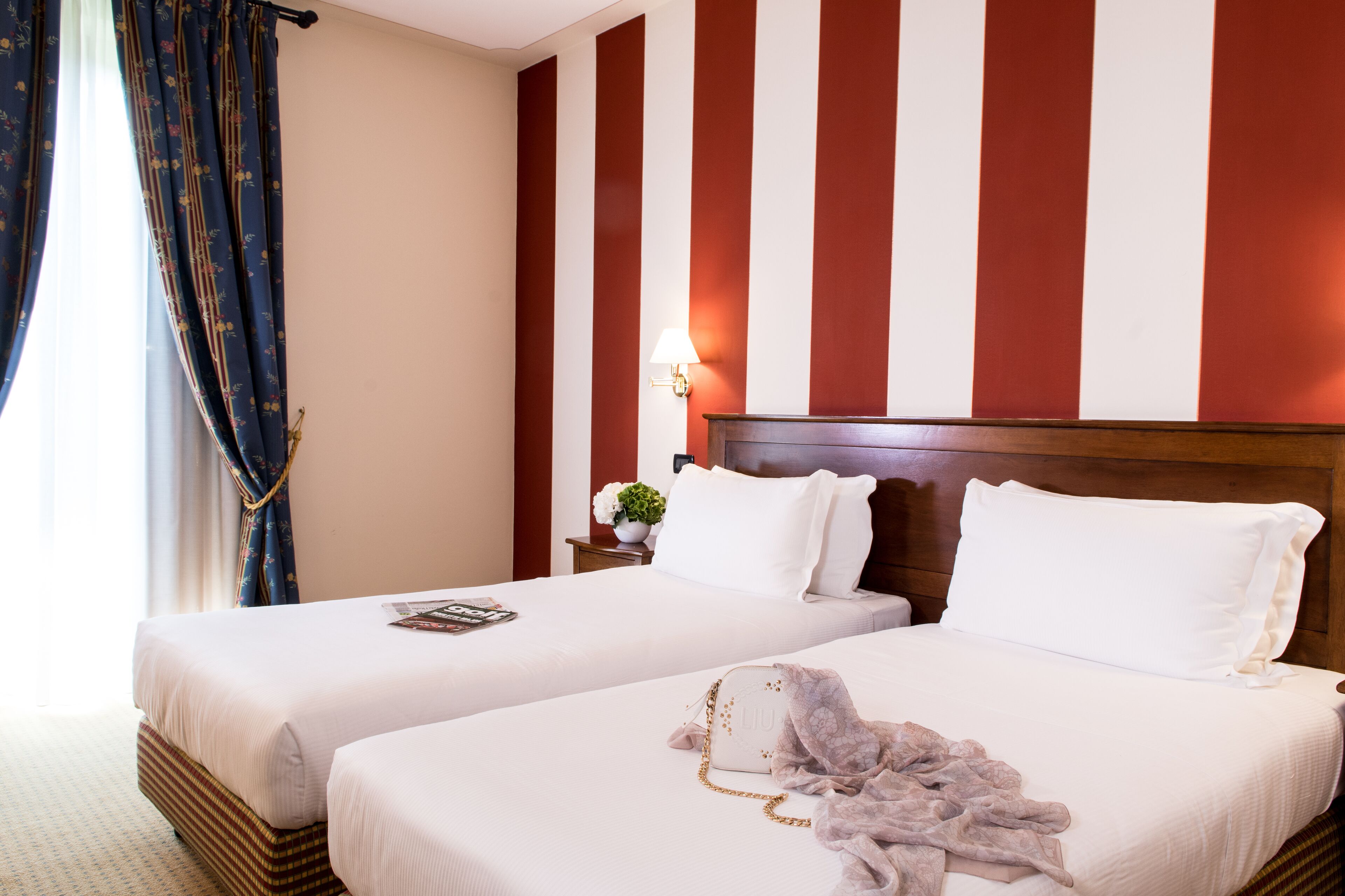 Una camera d'albergo con due letti, una testiera in legno, un comodino con una lampada e una finestra con tende.
