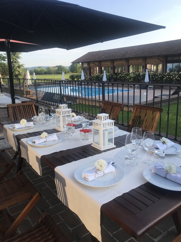 Outdoor dining tables set with white tablecloths, plates, and glasses, with a pool and garden in the background. - Relais Santo Stefano (Sandigliano)