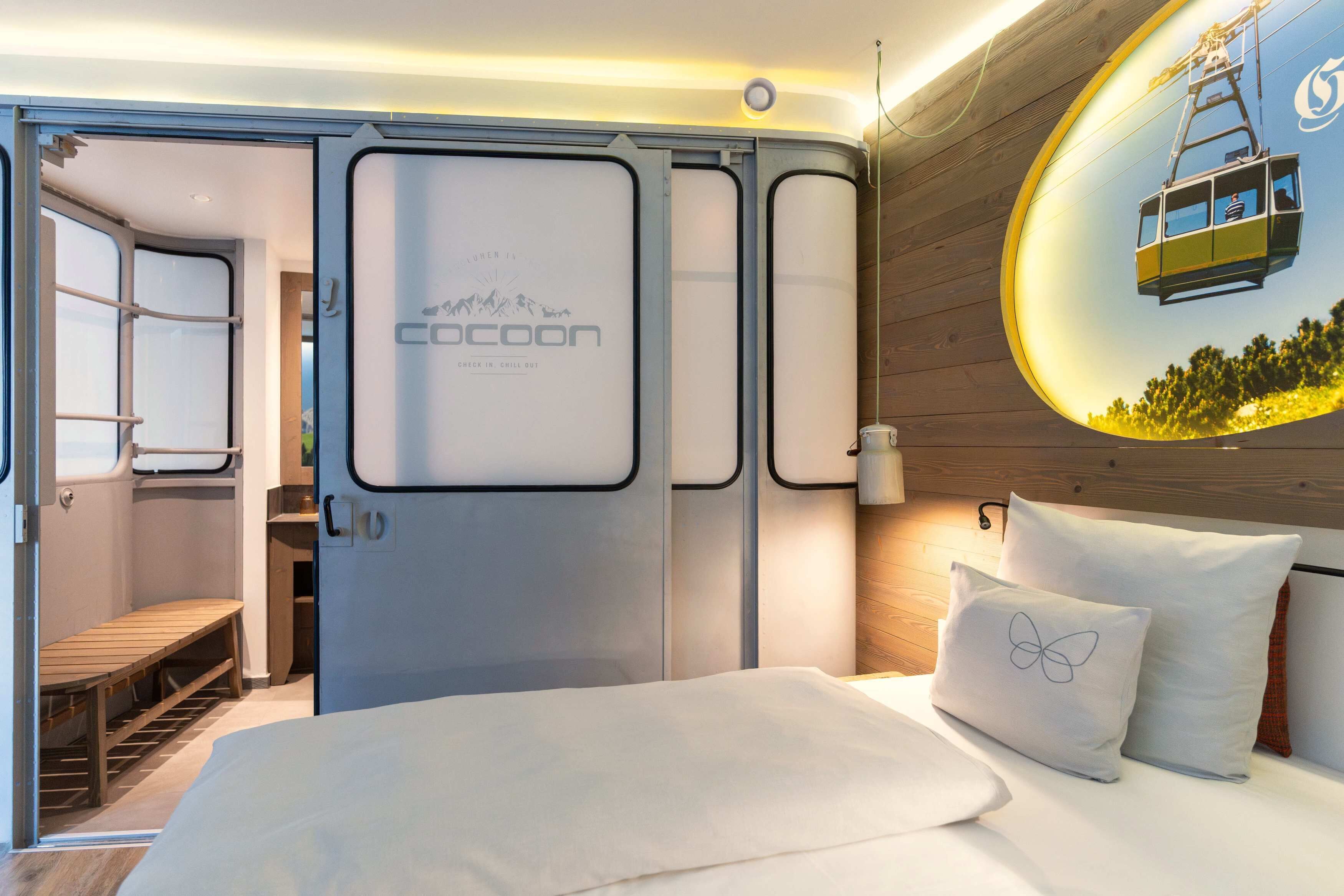 Een moderne hotelkamer met een bed, een houten hoofdeinde, een wandgemonteerde televisie en een raam met uitzicht op een kabelbaan.