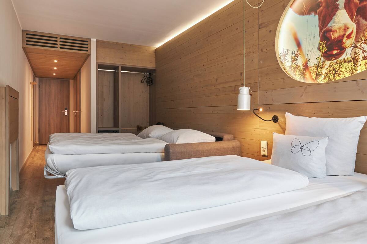 A hotel room with three beds, wooden walls, a ceiling vent, and a wall-mounted light fixture.