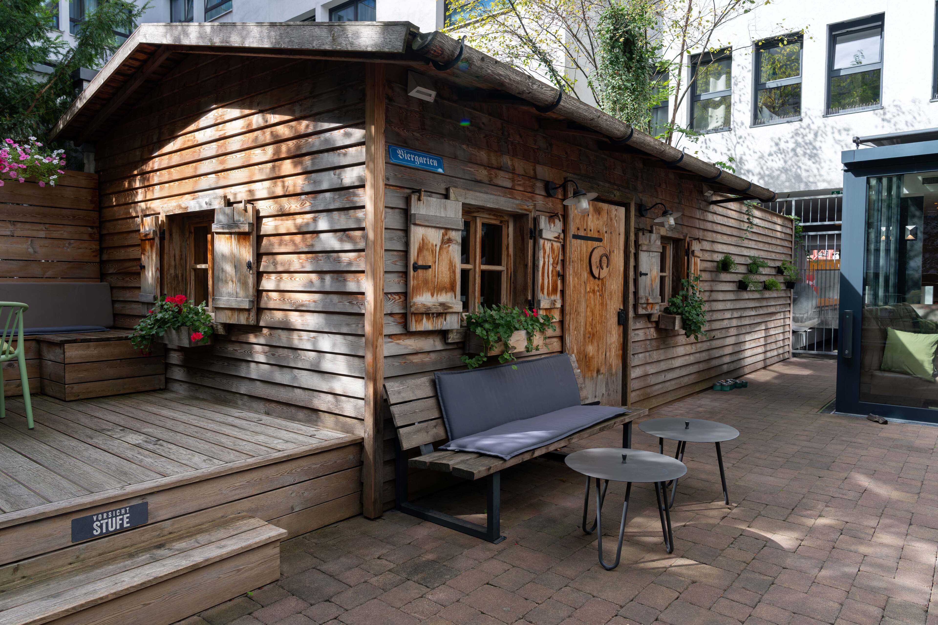 Une cabane en bois avec un banc et deux petites tables, une enseigne indiquant « Biergarten » et une autre signalant la présence d’une marche : « Vorsicht Stufe » (attention à la marche).
