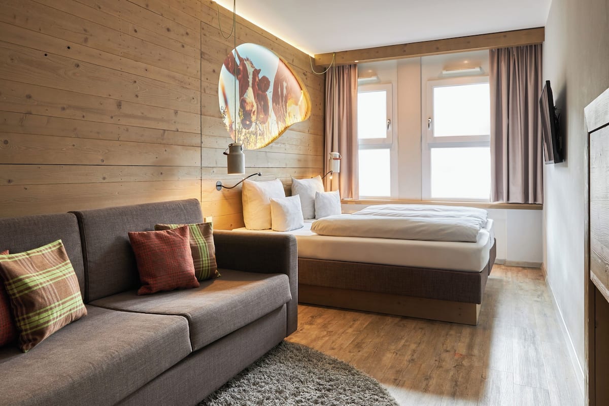 A modern hotel room with a wooden wall, a sofa, a bed, and a large window with curtains.